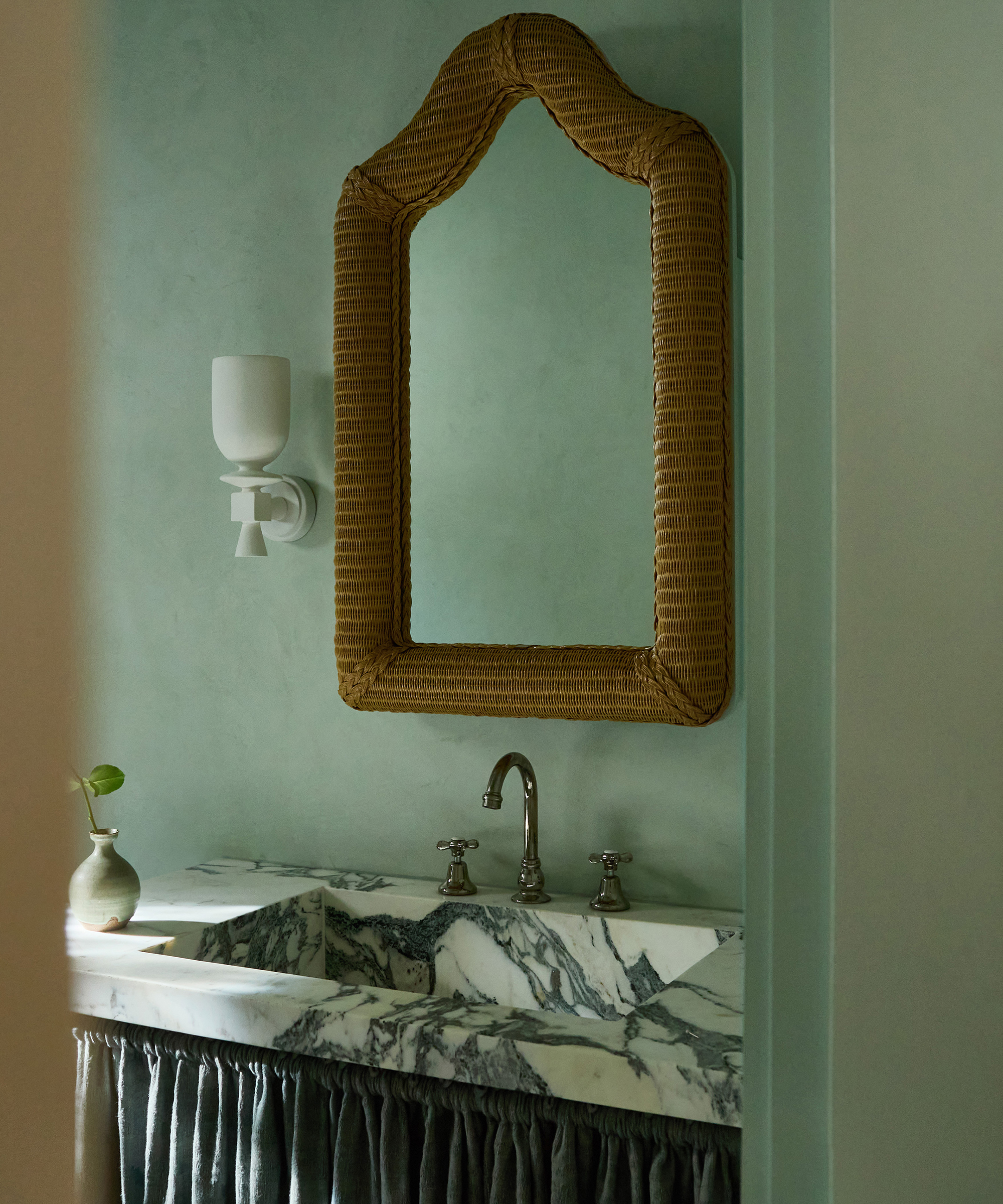 A bathroom in a Spanish style home with a marble sink, silver faucet, ornate wicker mirror, wall-mounted white sconce, and a small vase with a green leaf, set against pale green walls and a pleated fabric sink skirt.