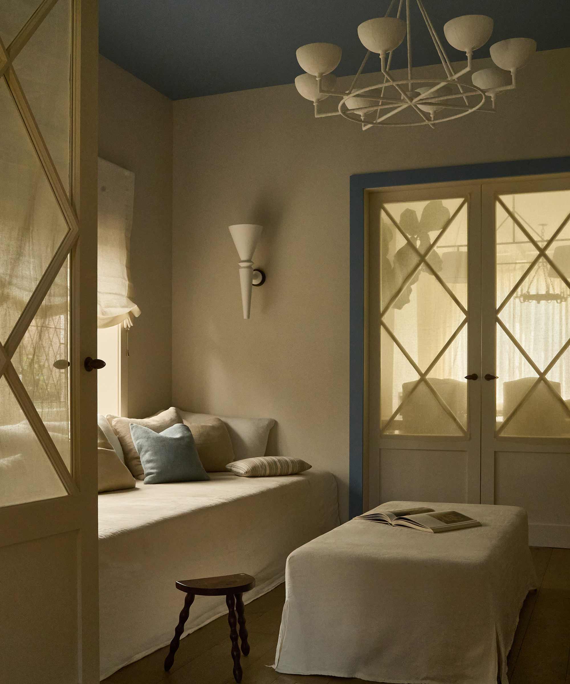 A cozy, softly-lit room in a Spanish style home with a white sofa, blue and beige pillows, a small wooden stool, and a fabric-covered ottoman with an open book. Light filters through glass doors with pale curtains and a modern chandelier above.