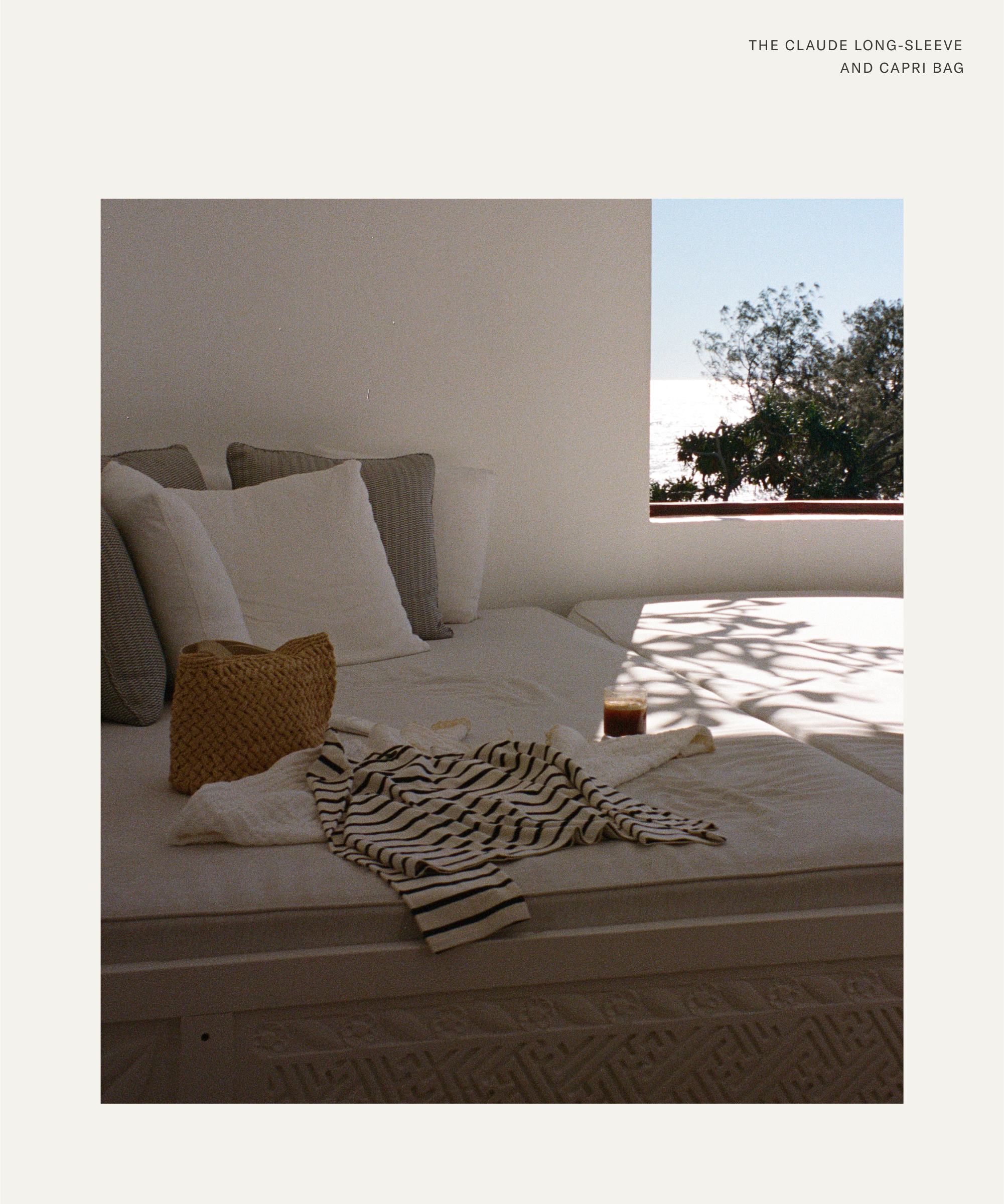 A cozy bed with white and gray pillows sits by a sunlit window. A striped shirt, Olive Cooke woven bag, and glass of iced coffee rest on the bed. Trees and a bright sky are visible outside.