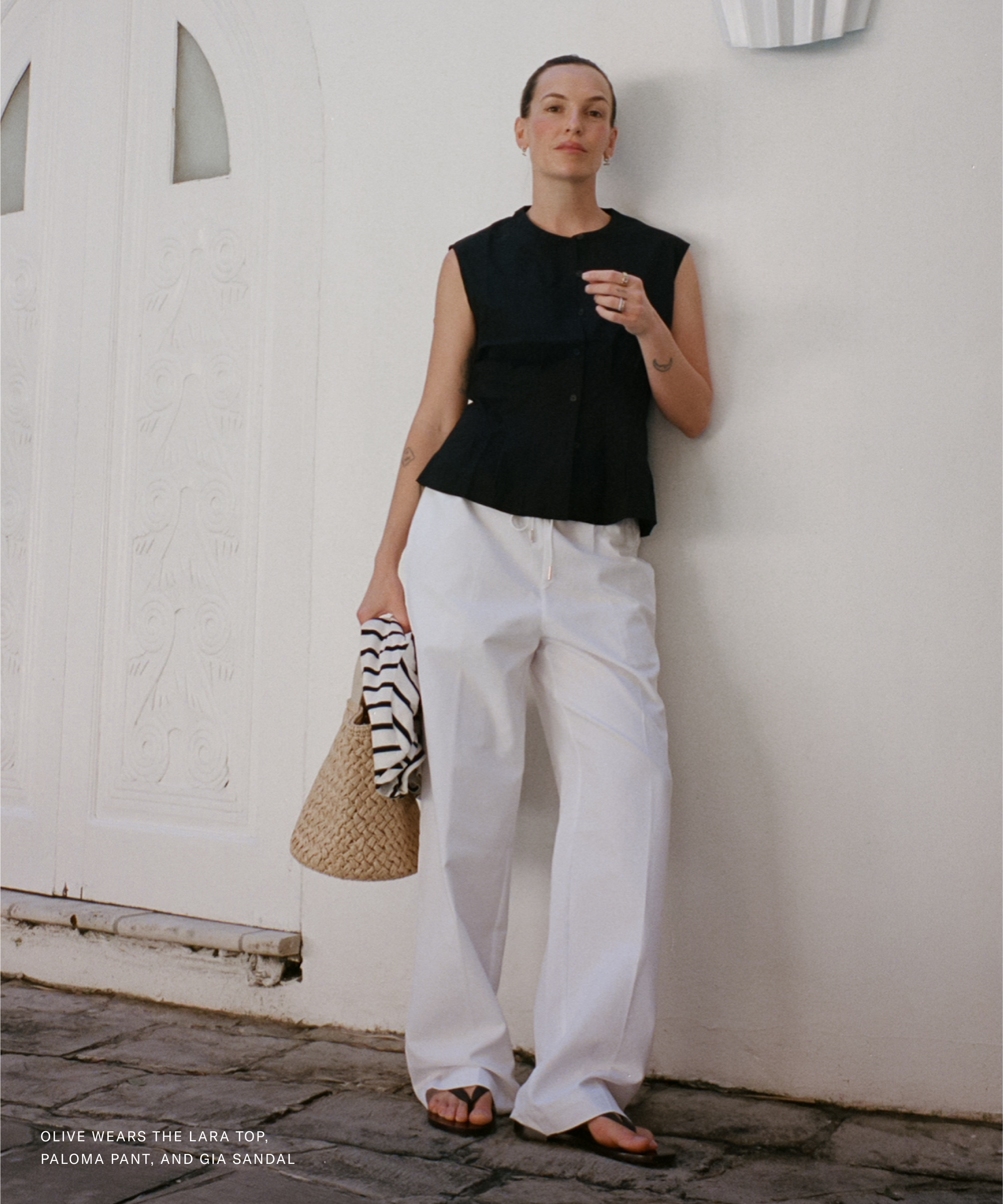 A woman stands by a white wall and ornate door, channeling Olive Cooke in a sleeveless black top, loose white pants, and black sandals. She holds a striped shirt and straw bag, looking relaxed and stylish.