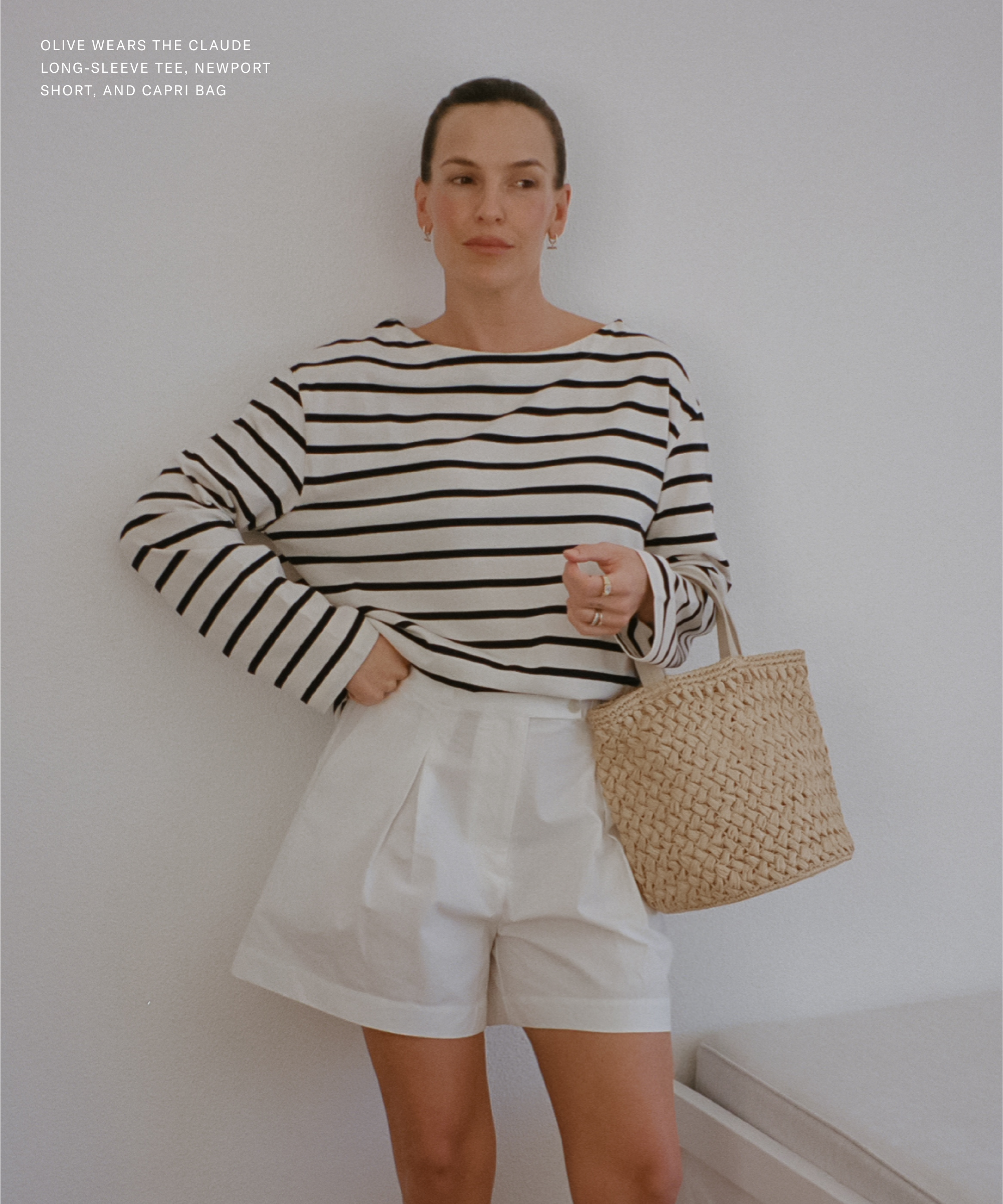 Channeling Olive Cookes effortless style, a woman stands against a white wall in a black-and-white striped long-sleeve tee, white shorts, and holding a woven straw bag. Her hair is pulled back and she radiates calm sophistication.