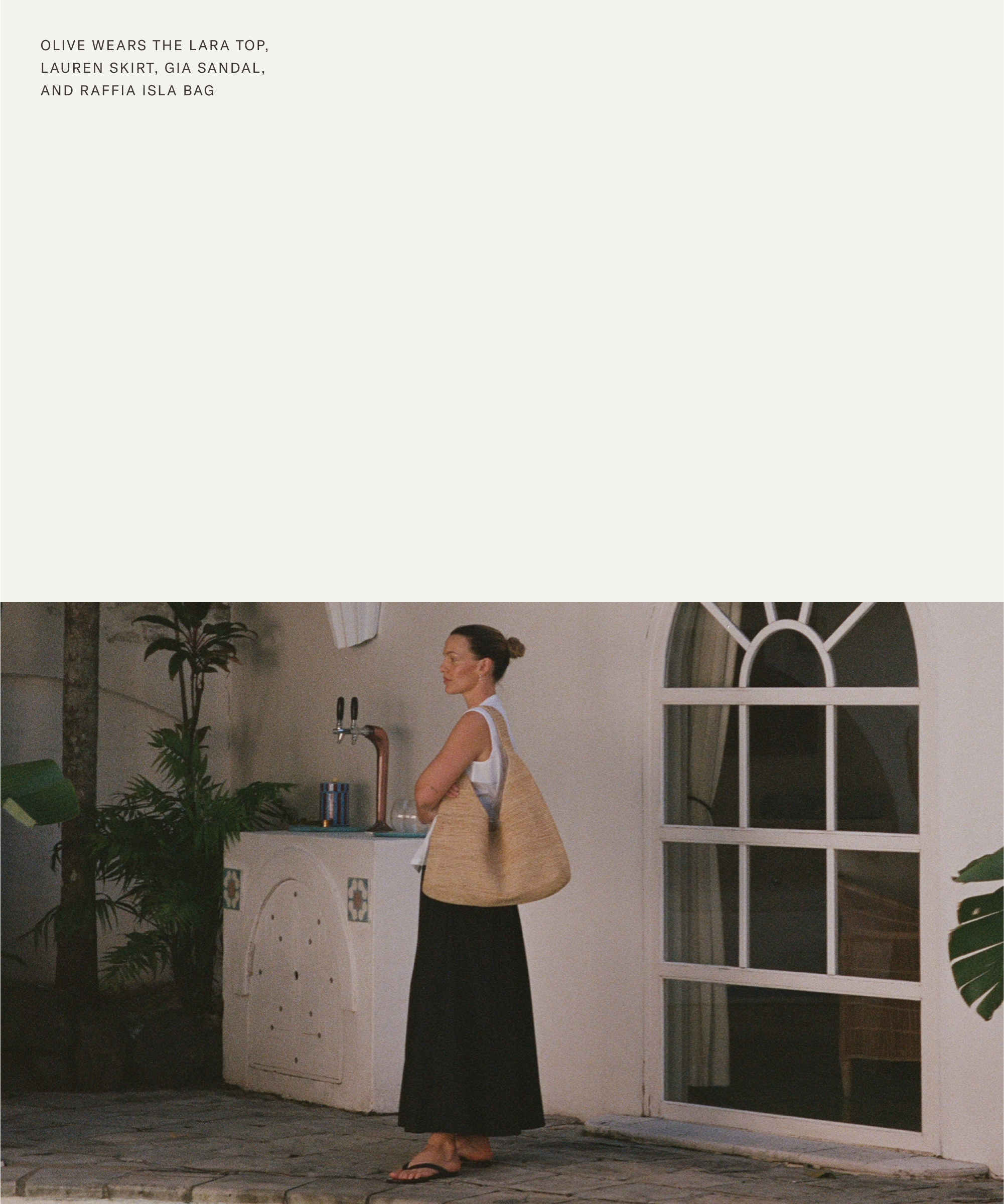 A woman, channeling Olive Cooke’s effortless style, stands outdoors by a white wall and window in a sleeveless white top, long black skirt, and black sandals, carrying a large raffia bag near a garden sink surrounded by green plants.