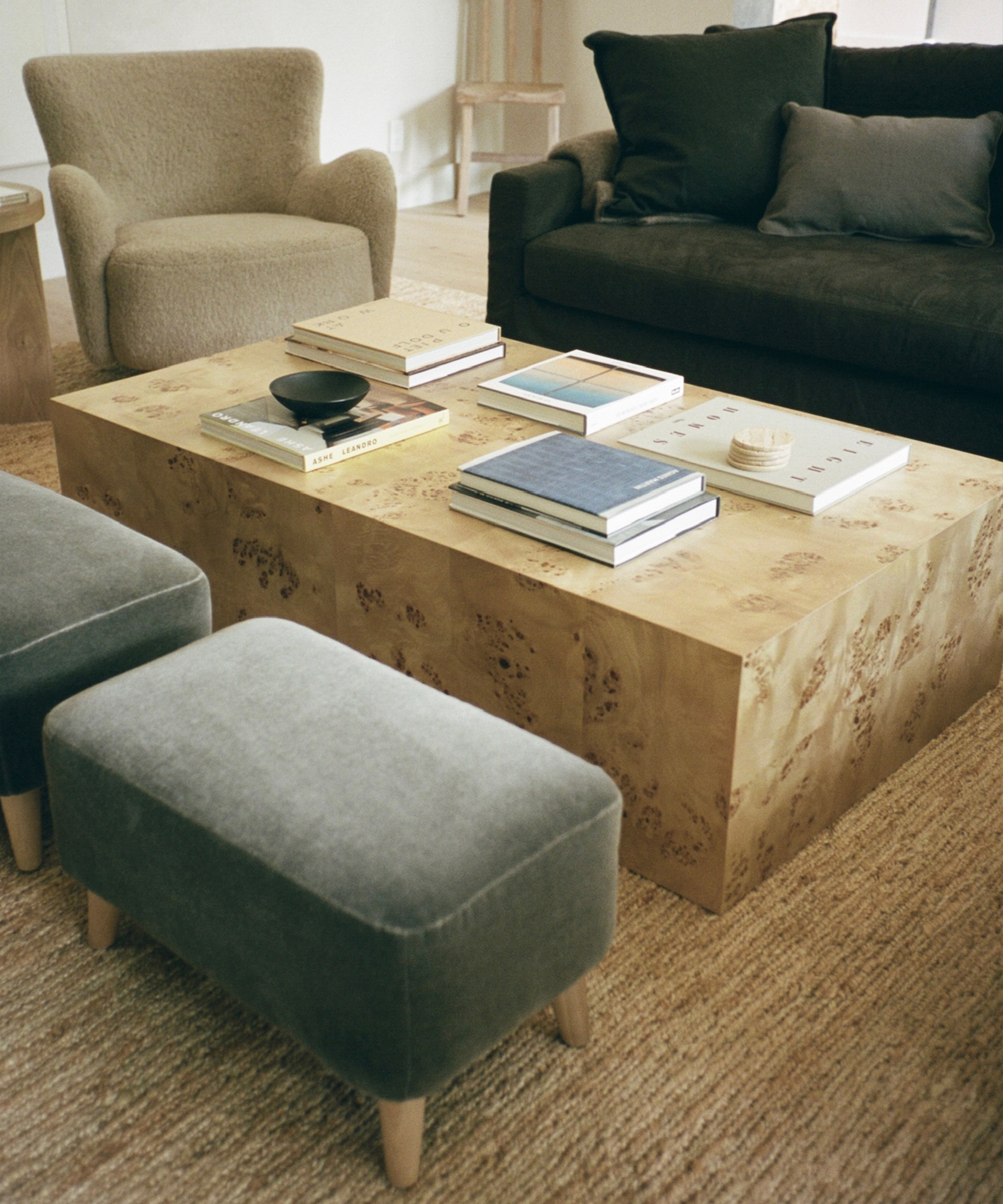 A modern living room shows how to style a beige textured coffee table with books and decor, paired with a dark sofa, a textured armchair, two gray ottomans, and a woven rug covering the floor.
