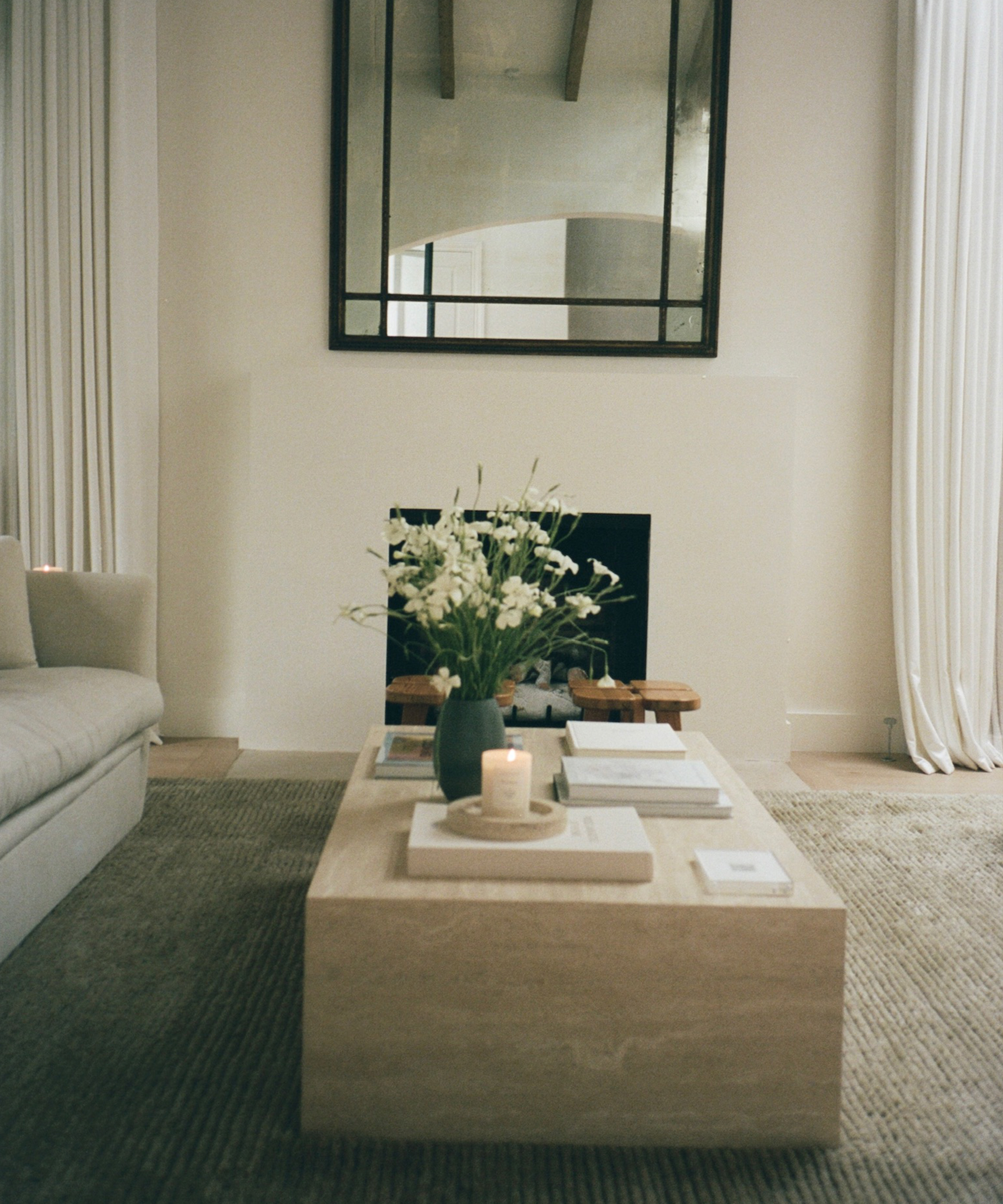 A minimalist living room with a beige sofa and light stone coffee table styled with books, a candle, and white flowers shows how to style serene spaces. Framed by light curtains, a fireplace with a large mirror completes the look.