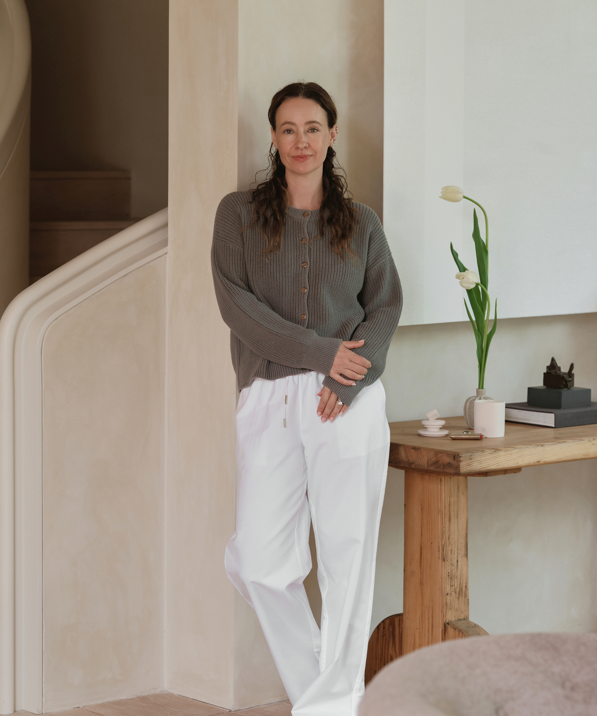 A woman with long brown hair, dressed in a gray sweater and white pants—perfect summer outfits—stands indoors, leaning against a wall beside a wooden table with a white vase holding a single tulip, books, and a cup.