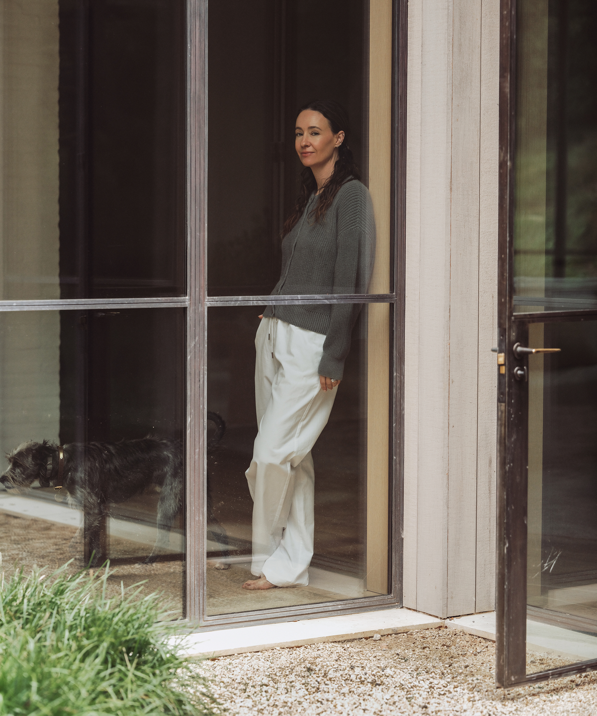 A woman in a gray sweater and white pants stands barefoot inside near a large window, smiling. A black dog stands nearby. The scene is calm, with greenery and gravel outside—a peaceful take on relaxed summer outfits.