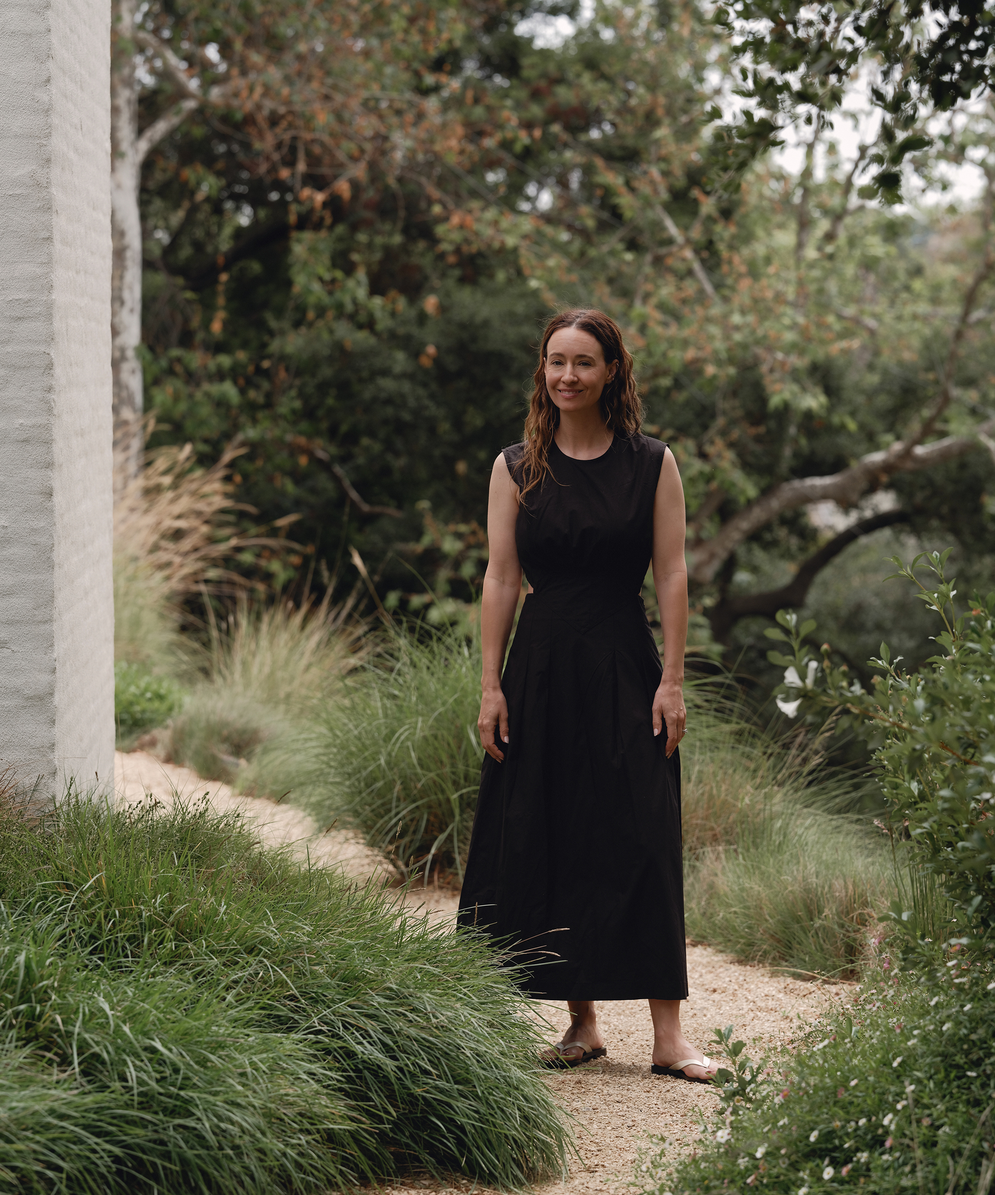 A woman in a sleeveless black dress, perfect for summer outfits, stands barefoot on a gravel pathway surrounded by tall grass and greenery, with trees and a white brick wall in the background.