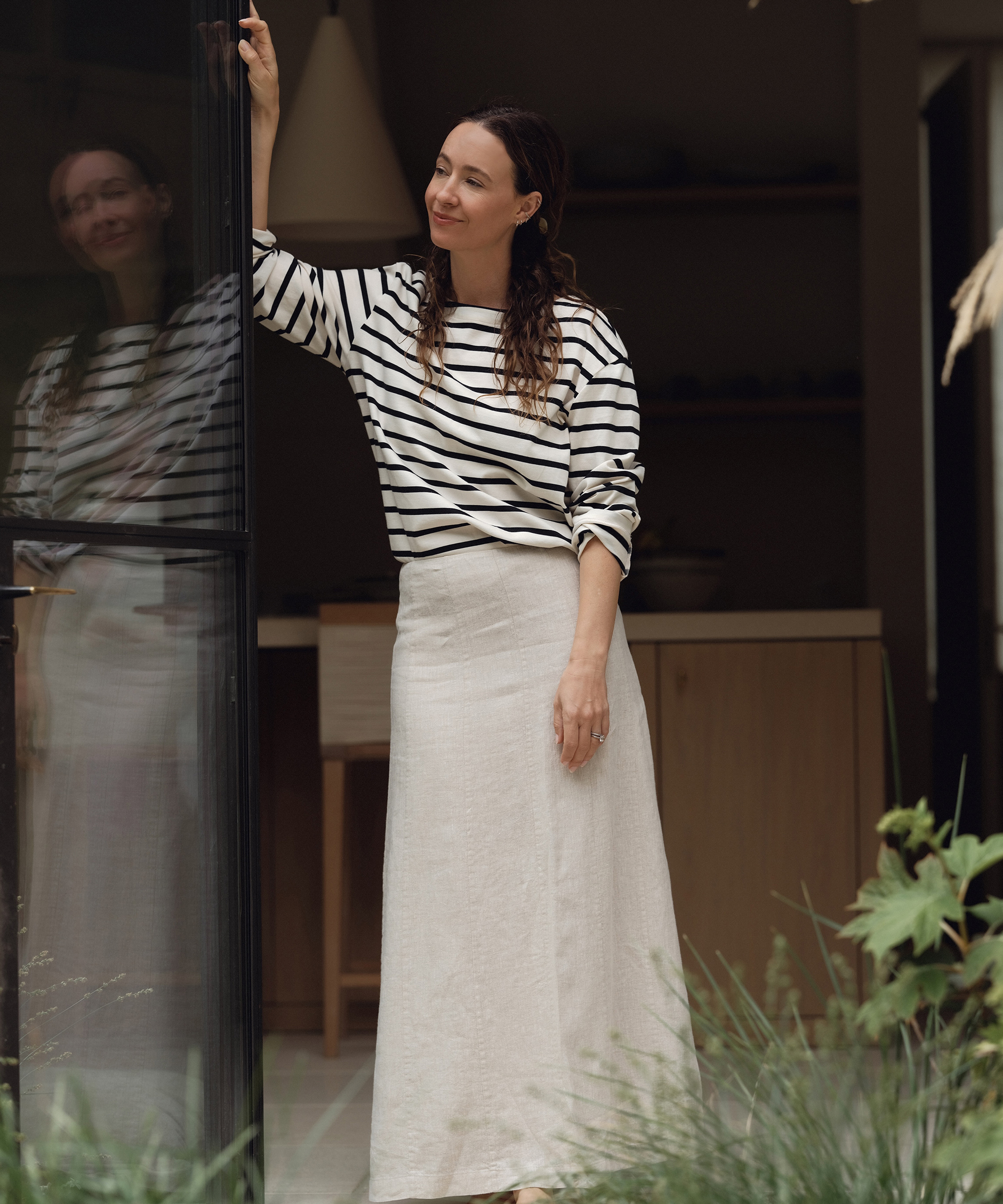 A woman in a striped long-sleeve shirt and a long white skirt stands by an open glass door, lightly touching the frame, her reflection visible in the glass. She is smiling and looking into the distance, embodying effortless summer outfits style.