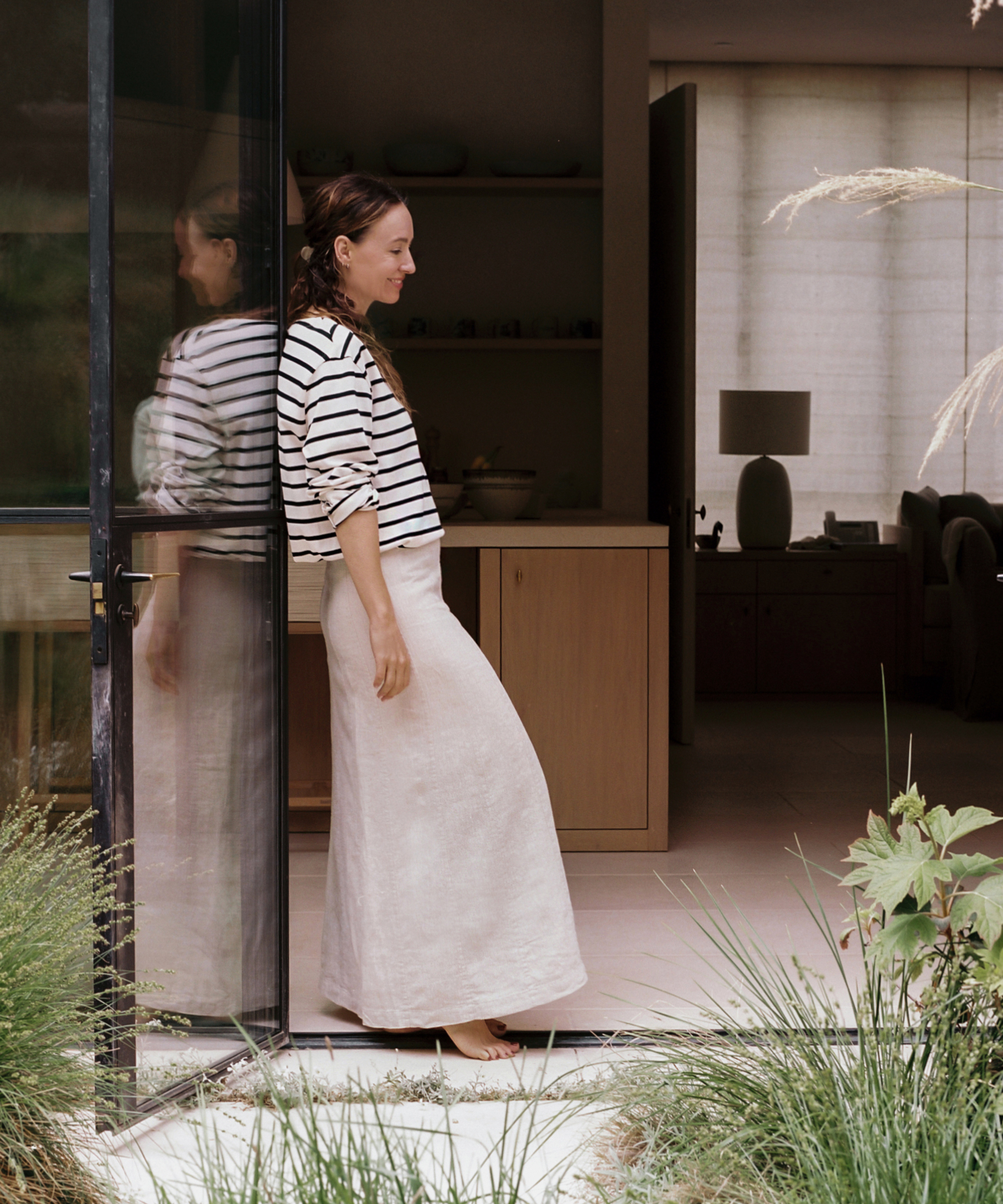 A woman in a striped shirt and long skirt leans against a glass door, smiling and looking down, showcasing effortless summer outfits with greenery in the foreground and a modern interior behind her.