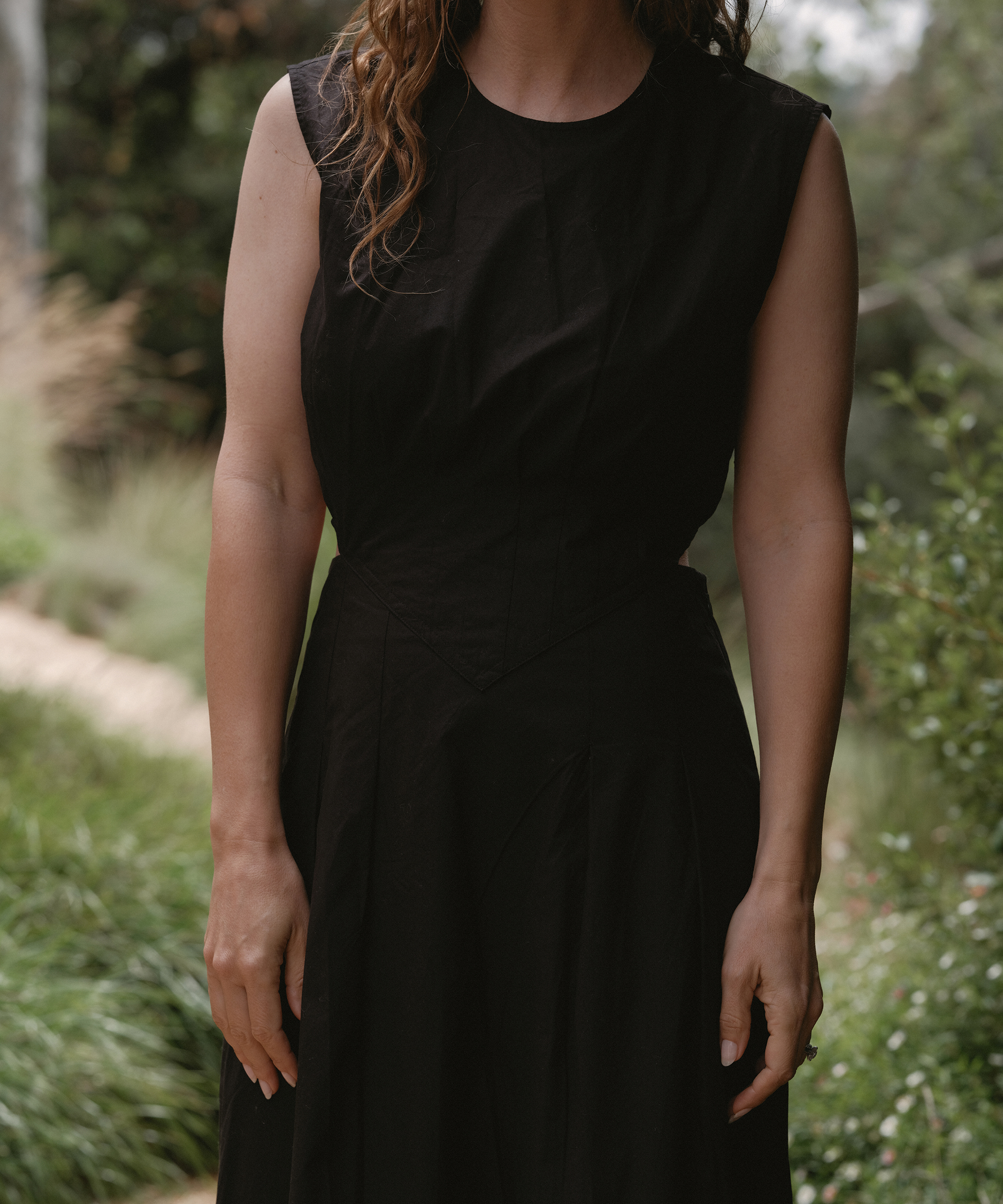 A woman with wavy brown hair wearing a sleeveless black dress, perfect for summer outfits, stands outdoors among green plants, her arms relaxed at her sides. Her face is partially out of frame.
