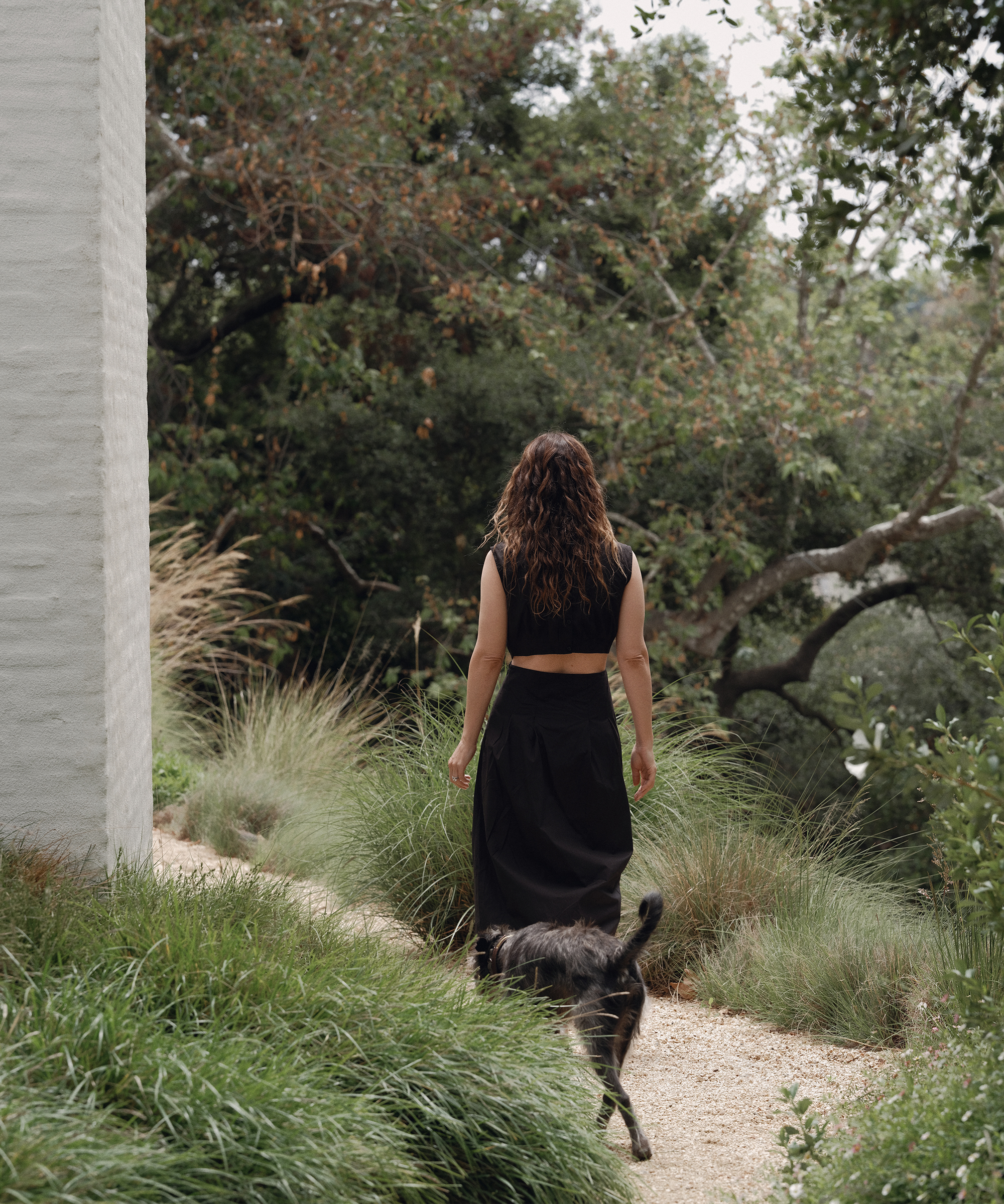 A woman in stylish summer outfits—with wavy brown hair, a black sleeveless top and skirt—walks along a garden path beside a white building, accompanied by her black dog. Lush greenery and trees surround them.