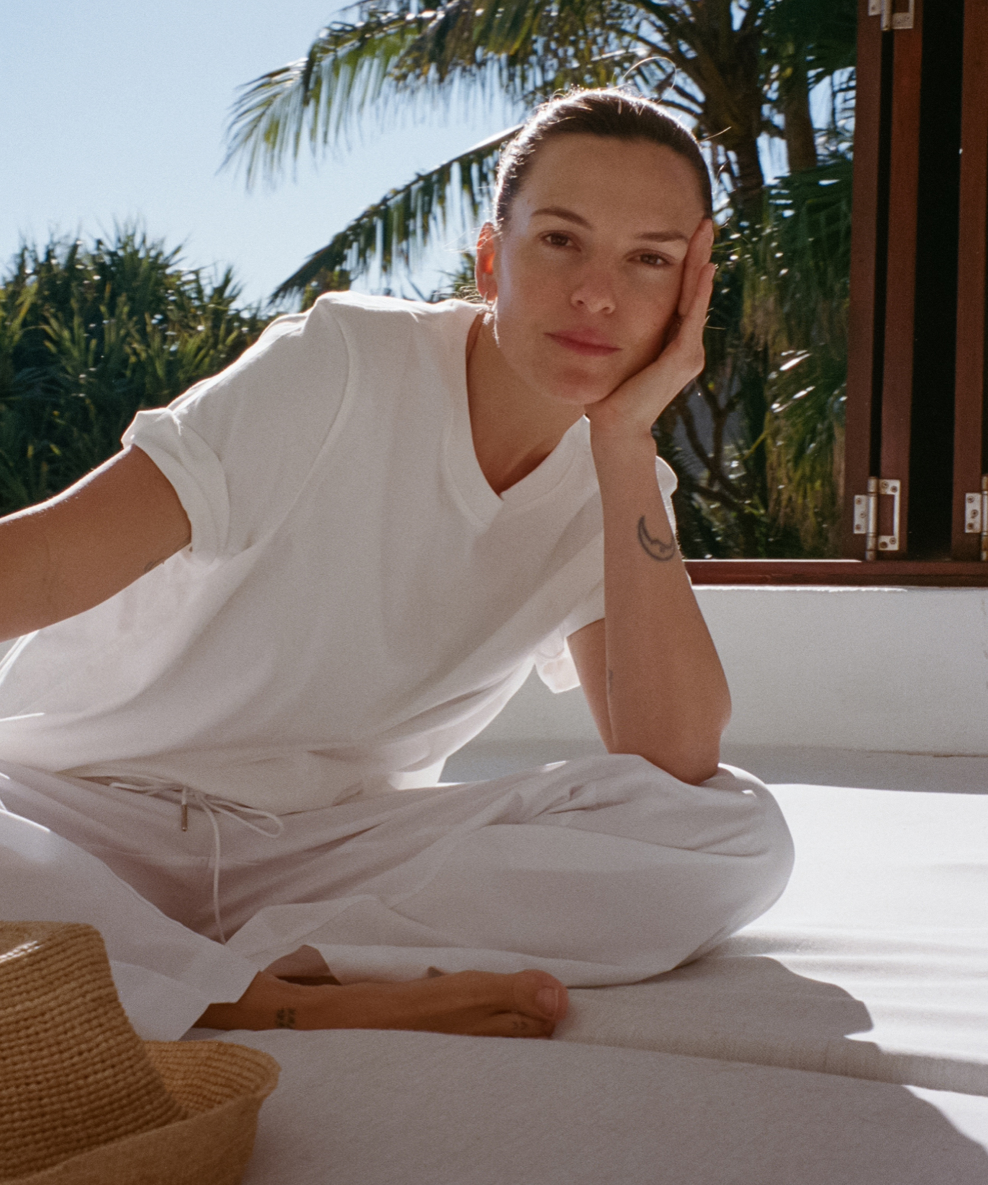olive cooke wearing a white tee and white pants sitting outside on a white cushion