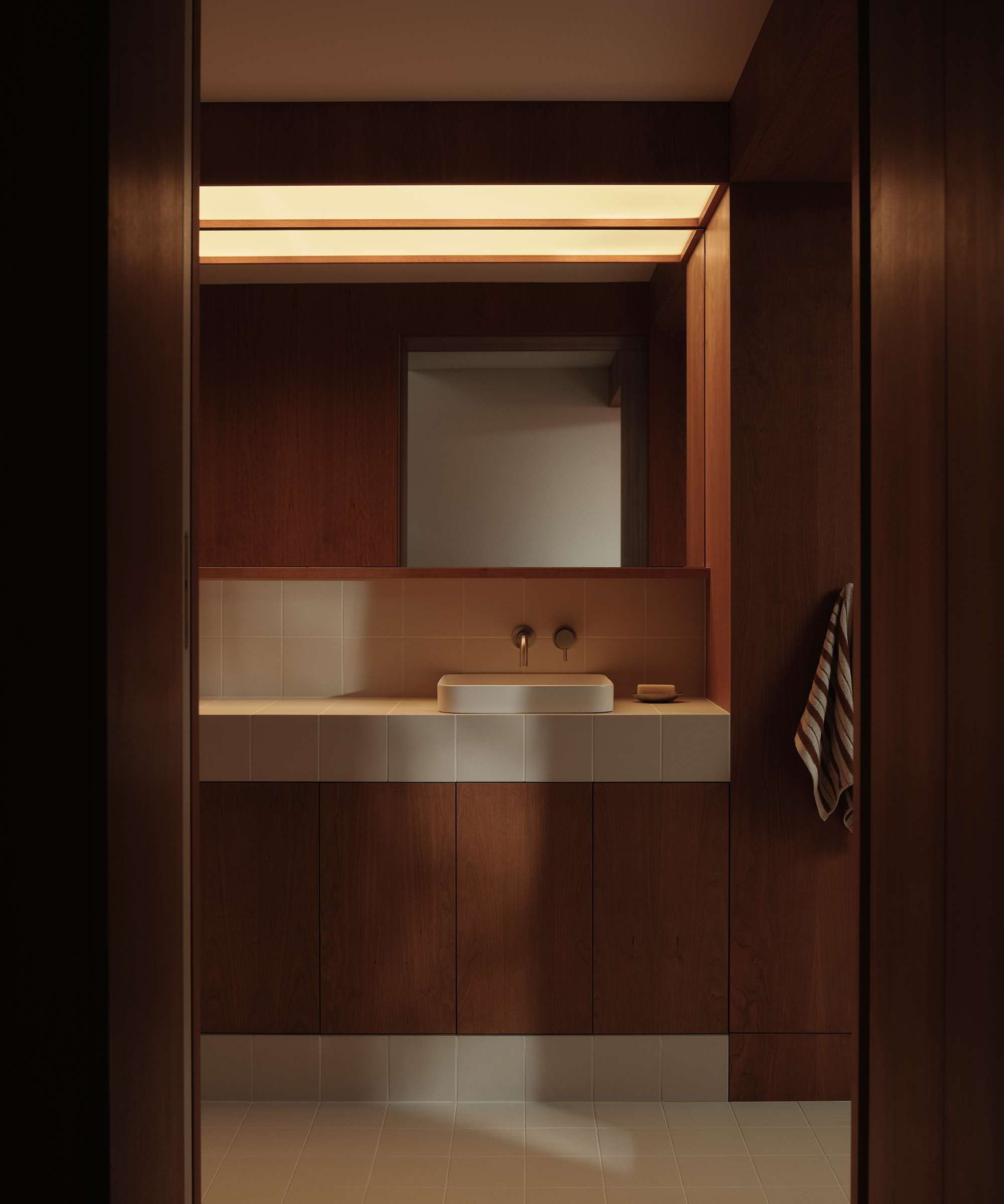 A minimalist bathroom with wood-paneled walls and midcentury flair, featuring a white tiled counter with an integrated sink, a wall-mounted faucet, a mirror above the sink, and a striped towel. Warm lighting fills the space.