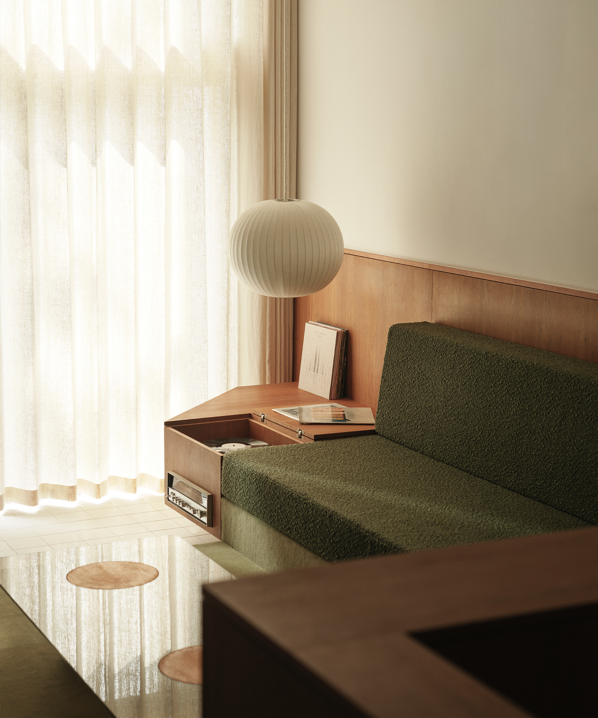 Minimalist living room with soft natural light, featuring a midcentury green upholstered sofa, a wooden side table with books, a round white hanging lamp, sheer curtains, and a glass coffee table with wooden accents.