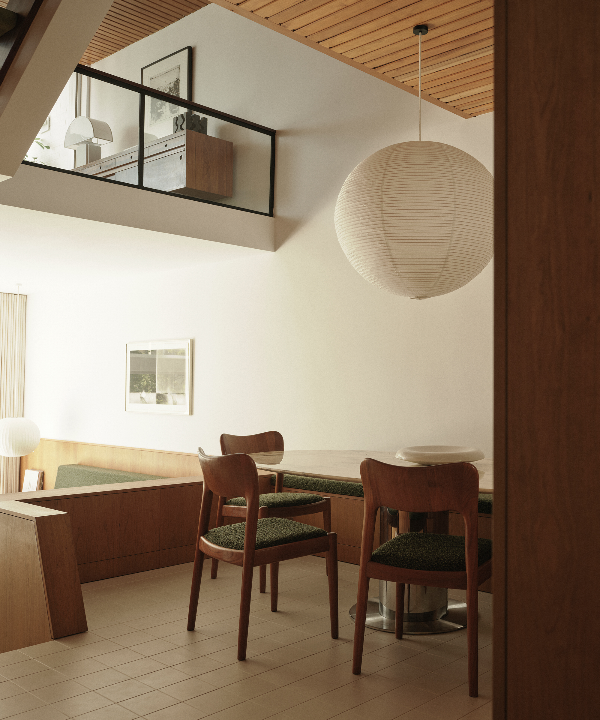 A modern, midcentury-inspired dining area with wooden chairs and a round table, green cushions, a large paper lantern light, wood accents, and a view of an upper level with glass railing and minimalist decor.