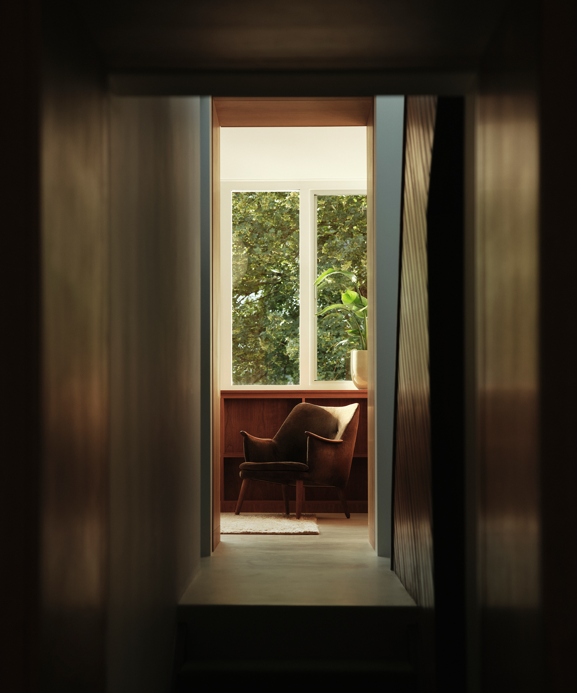 A cozy midcentury brown armchair sits by a window with sunlight streaming in, framed by greenery outside. The scene is viewed from a dim hallway, creating a warm and inviting nook.