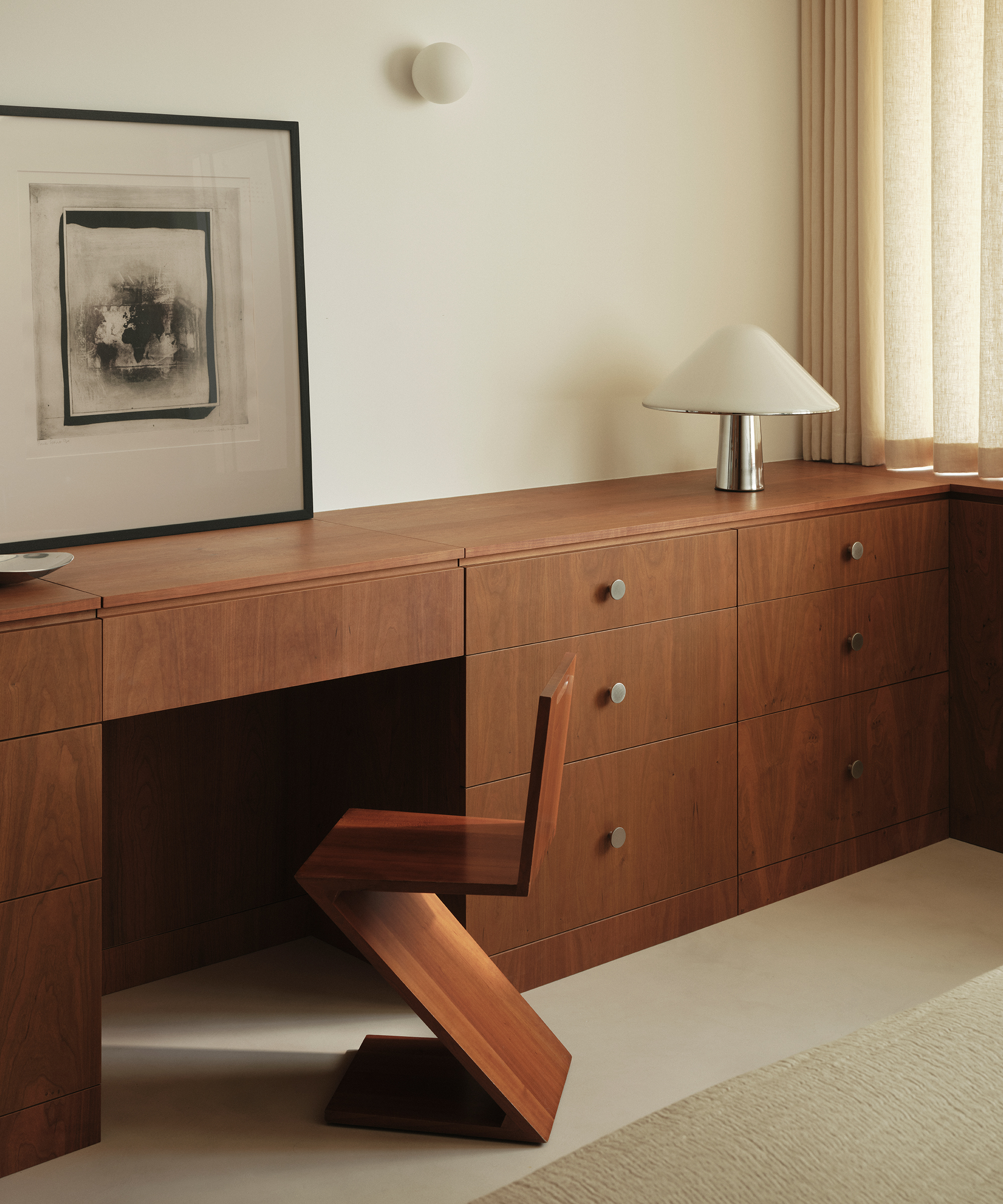 A minimalist midcentury workspace with a wooden desk and matching geometric chair, built-in drawers, a modern table lamp, abstract wall art, and beige curtains letting in natural light.