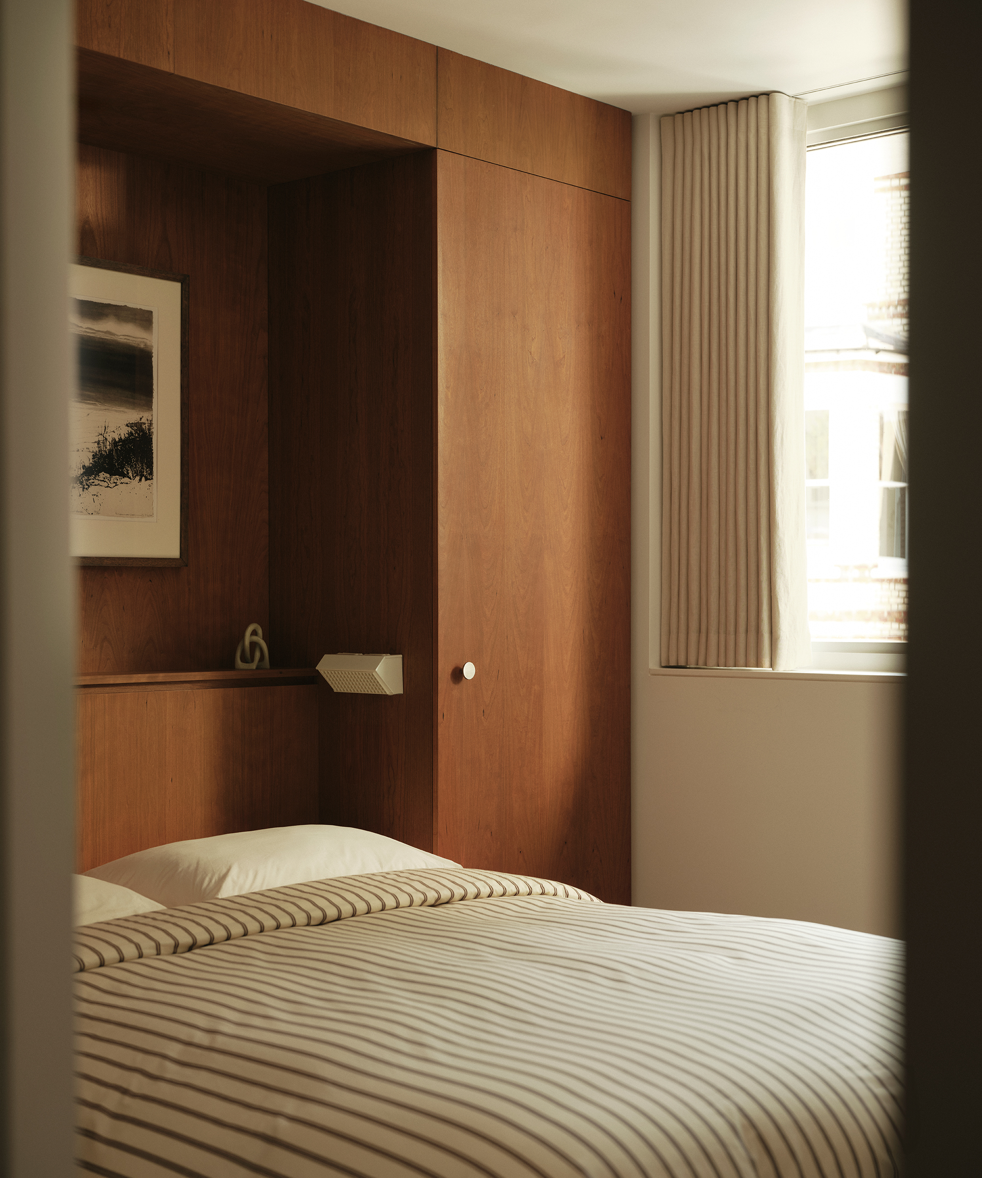 A neatly made bed with striped bedding in a cozy, modern room featuring midcentury wooden wall panels, a framed picture, and a window with cream curtains letting in natural light.