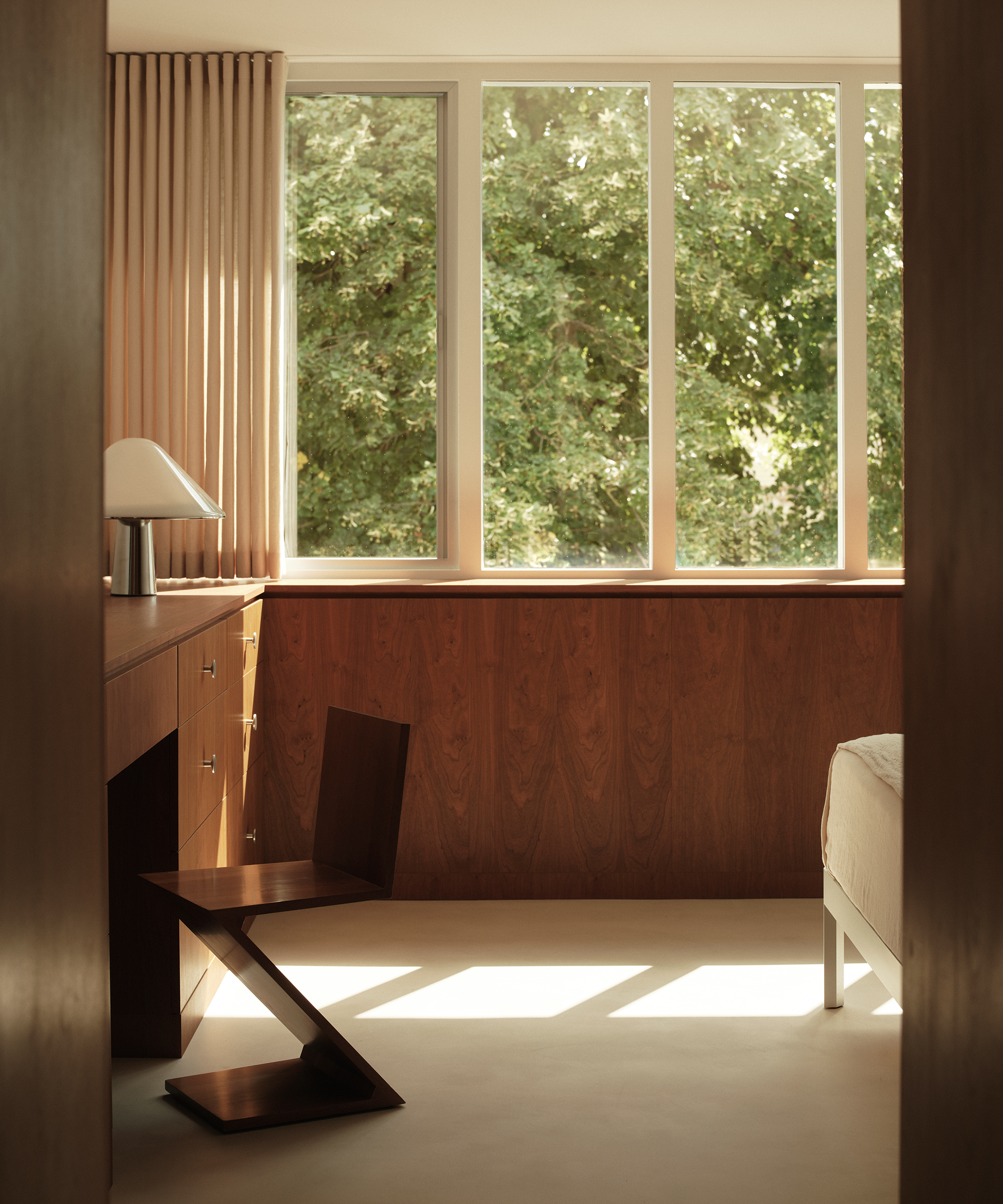 A minimalist room features a sculptural midcentury wooden chair at a wooden desk, next to a bed. Large windows let in natural light, revealing lush green trees outside. Neutral tones and soft sunlight create a calm, modern atmosphere.