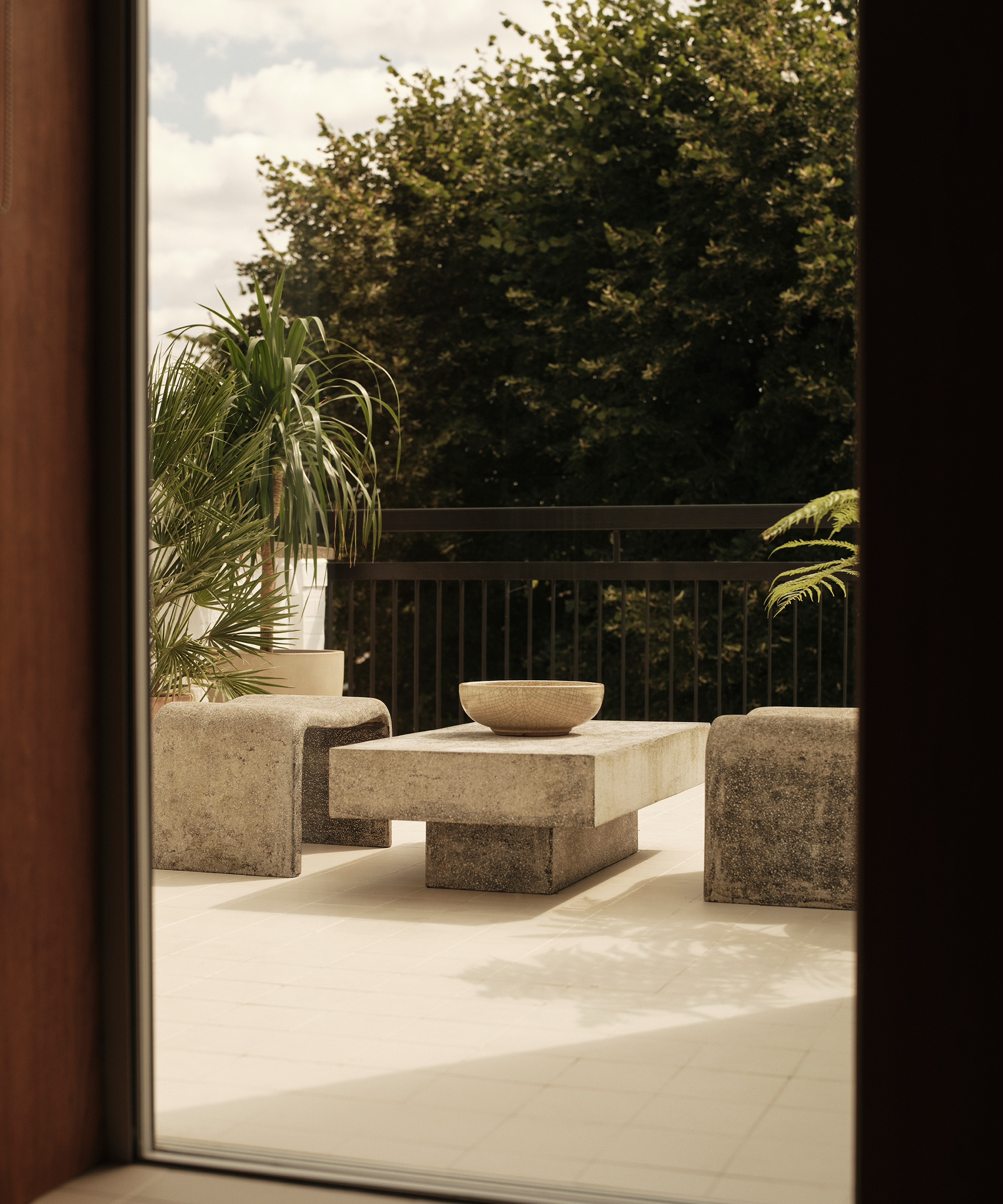 Concrete benches and a table with a bowl sit on a tiled outdoor patio, surrounded by potted plants and greenery, as seen through a window. The bright, serene setting evokes midcentury charm.