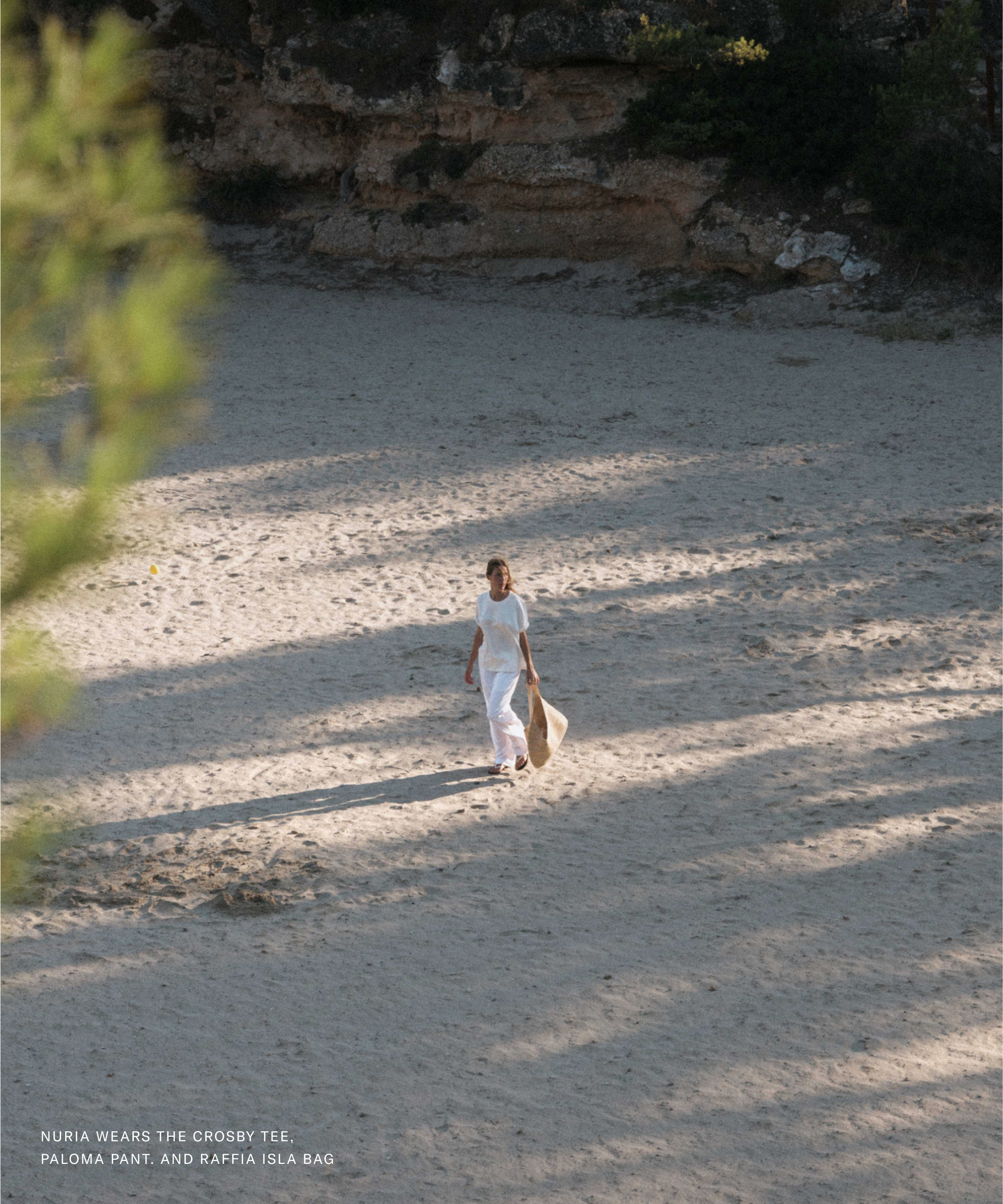 A person in a white outfit walks alone on a sandy area, carrying a light-colored bag. Shadows from nearby trees create patterns on the sand. Text at the bottom highlights Nuria Val’s clothes and accessories worn.