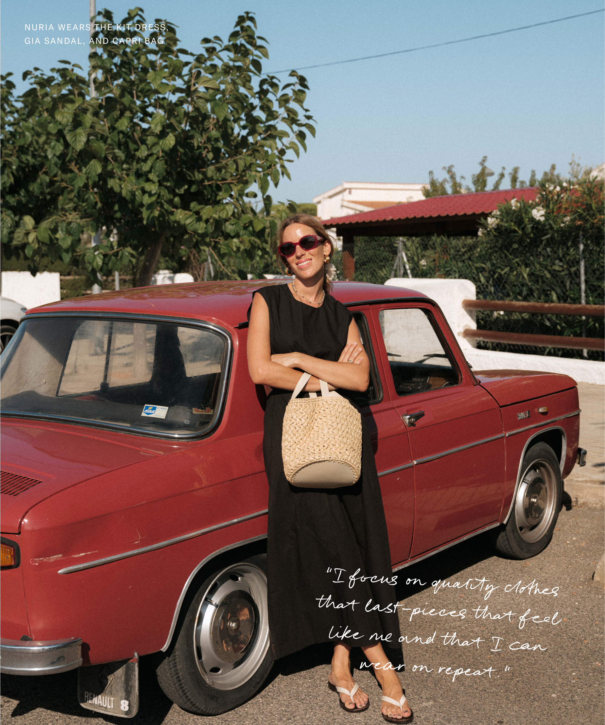 Nuria Val stands smiling in a sleeveless black dress and sandals beside a vintage red car, holding a straw bag. Trees and a white fence frame the sunny scene.