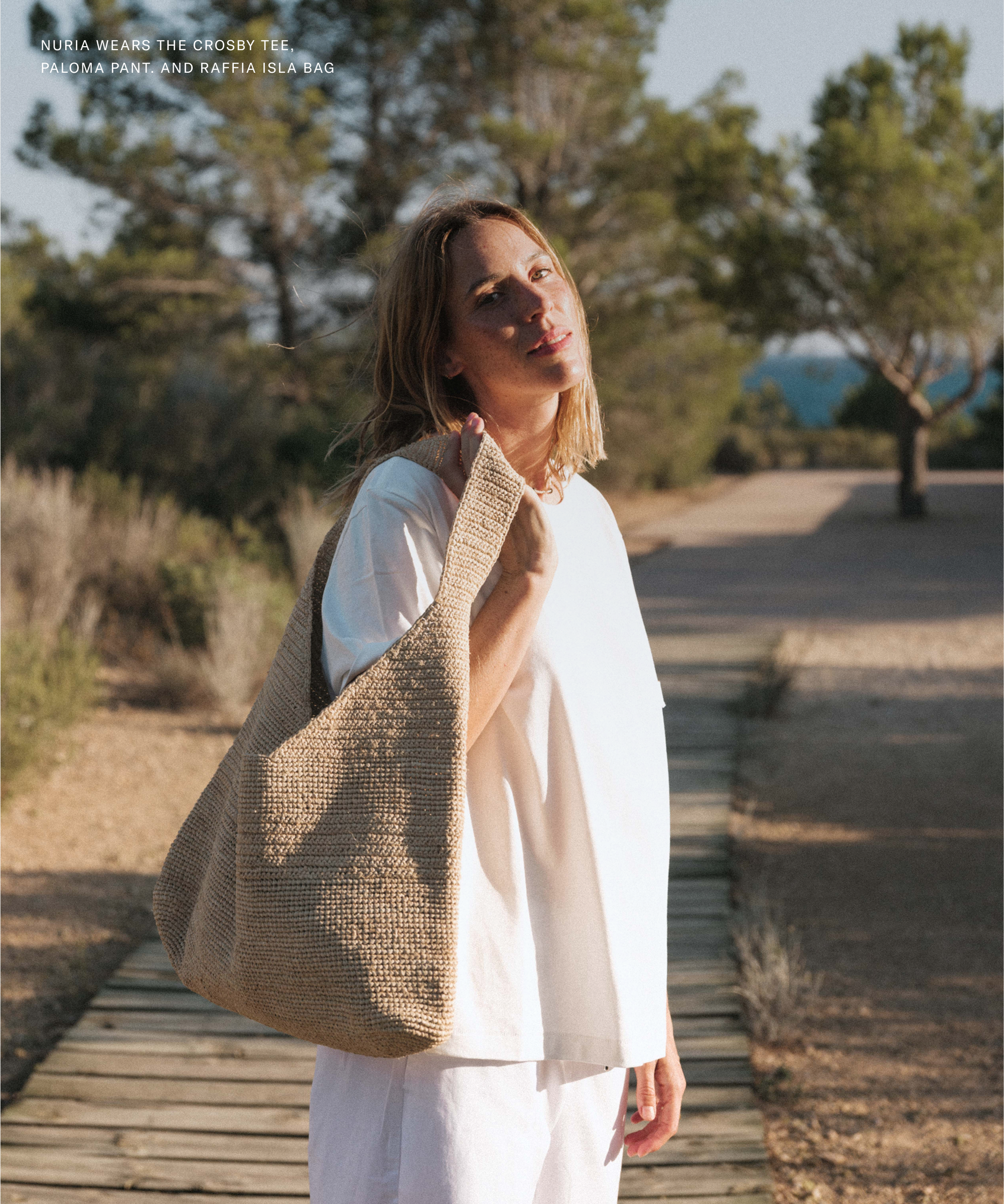 A woman stands on a sunlit wooden path outdoors, wearing a loose white t-shirt and pants, holding a large woven raffia bag over her shoulder. Inspired by Nuria Val, the scene features trees and blue sky in the background.