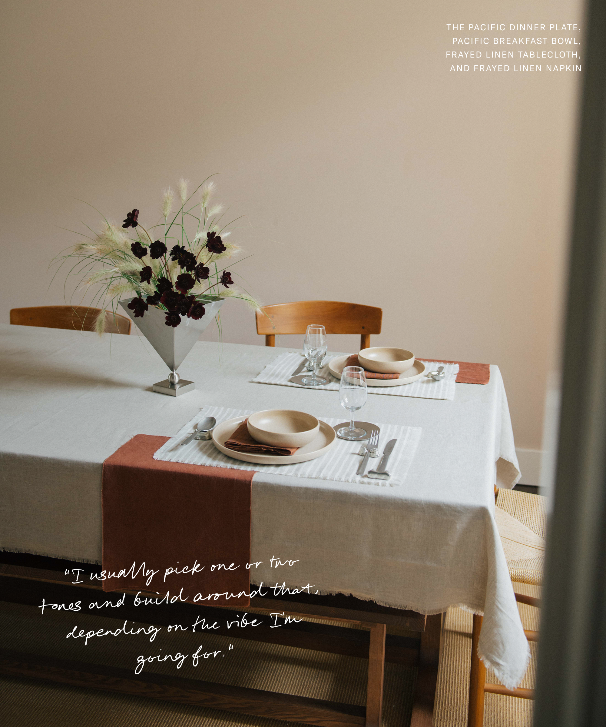 A dining table set by Kirthanaa Naidu features tan dishes, glassware, and a white tablecloth with a rust-colored runner. A vase with white and dark flowers centers the scene, flanked by two wooden chairs in soft natural lighting. Text is on the image.