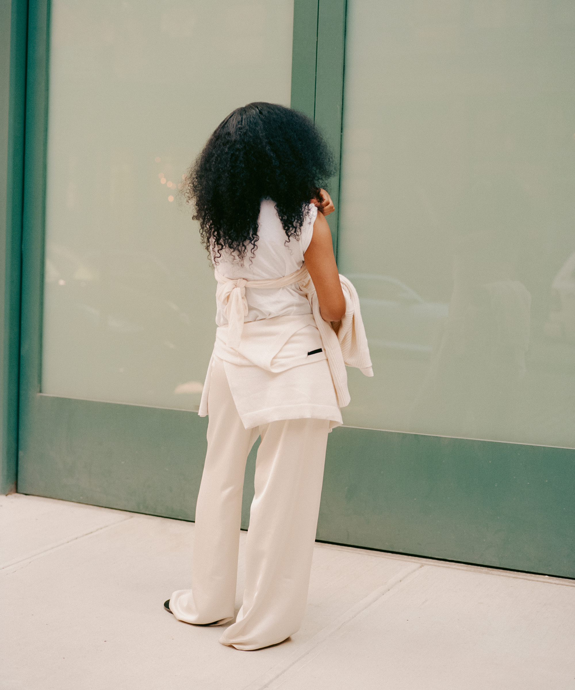 A woman with curly hair, dressed in loose-fitting cream-colored pants and a sleeveless top, stands on a sidewalk facing a frosted glass wall with a green frame, her back to the camera.
