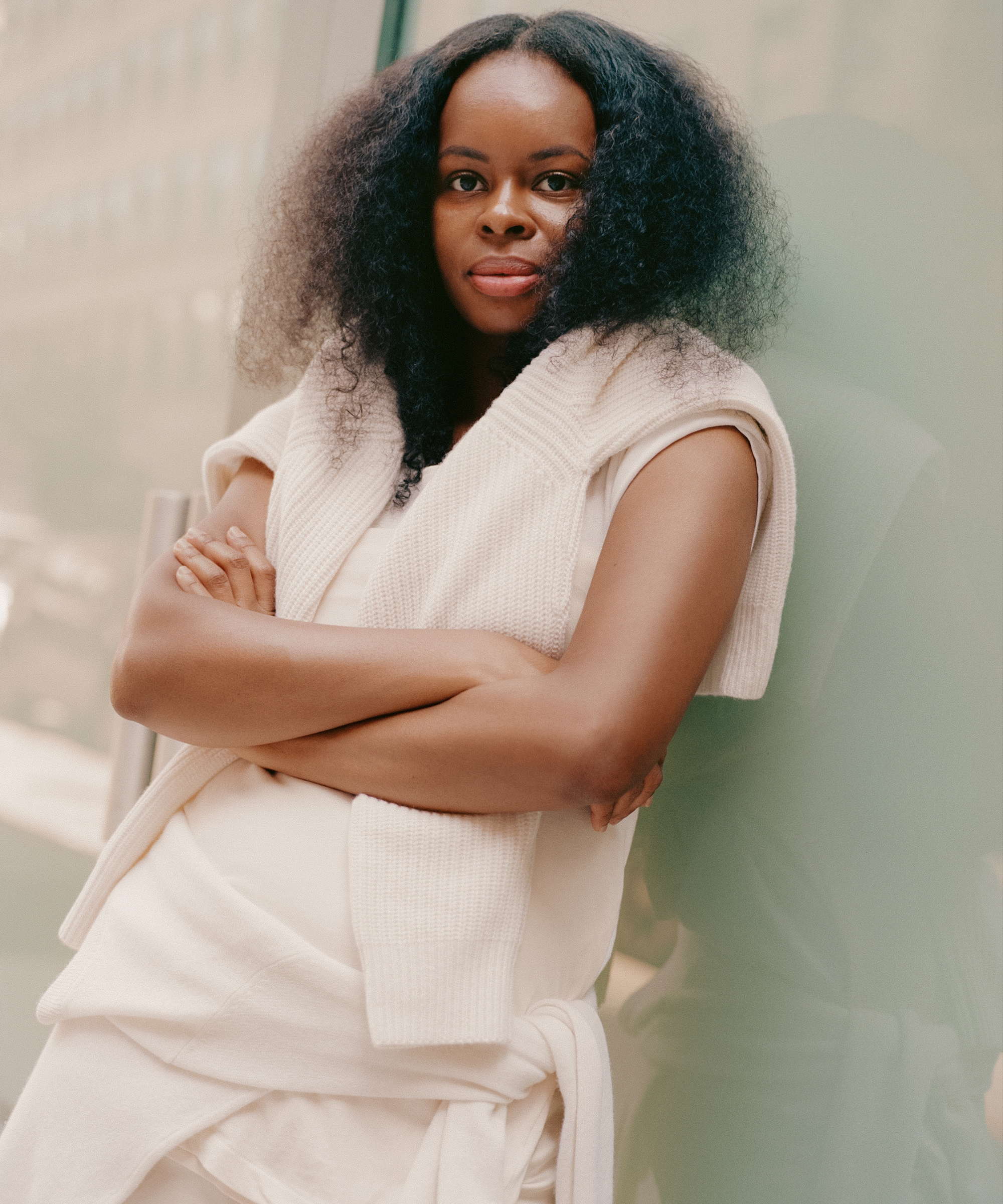 A woman with long, natural curly hair wearing a cream-colored outfit and sweater draped over her shoulders stands with arms crossed, leaning against a glass wall, looking confidently at the camera.