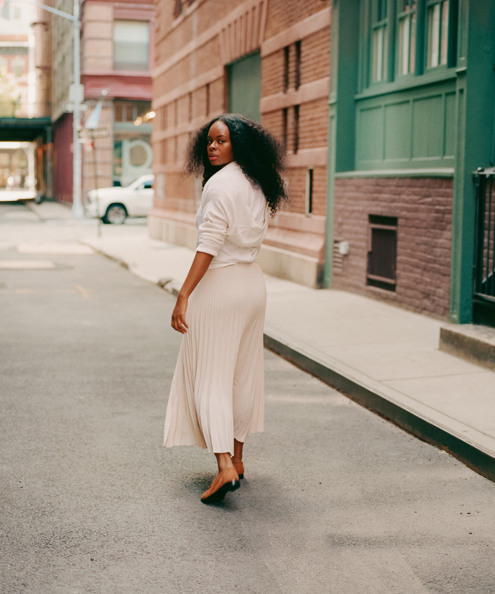 A woman with curly hair, wearing a white top and a flowing cream skirt, walks down a quiet city street, looking back over her shoulder. Brick buildings and a parked car are visible in the background.