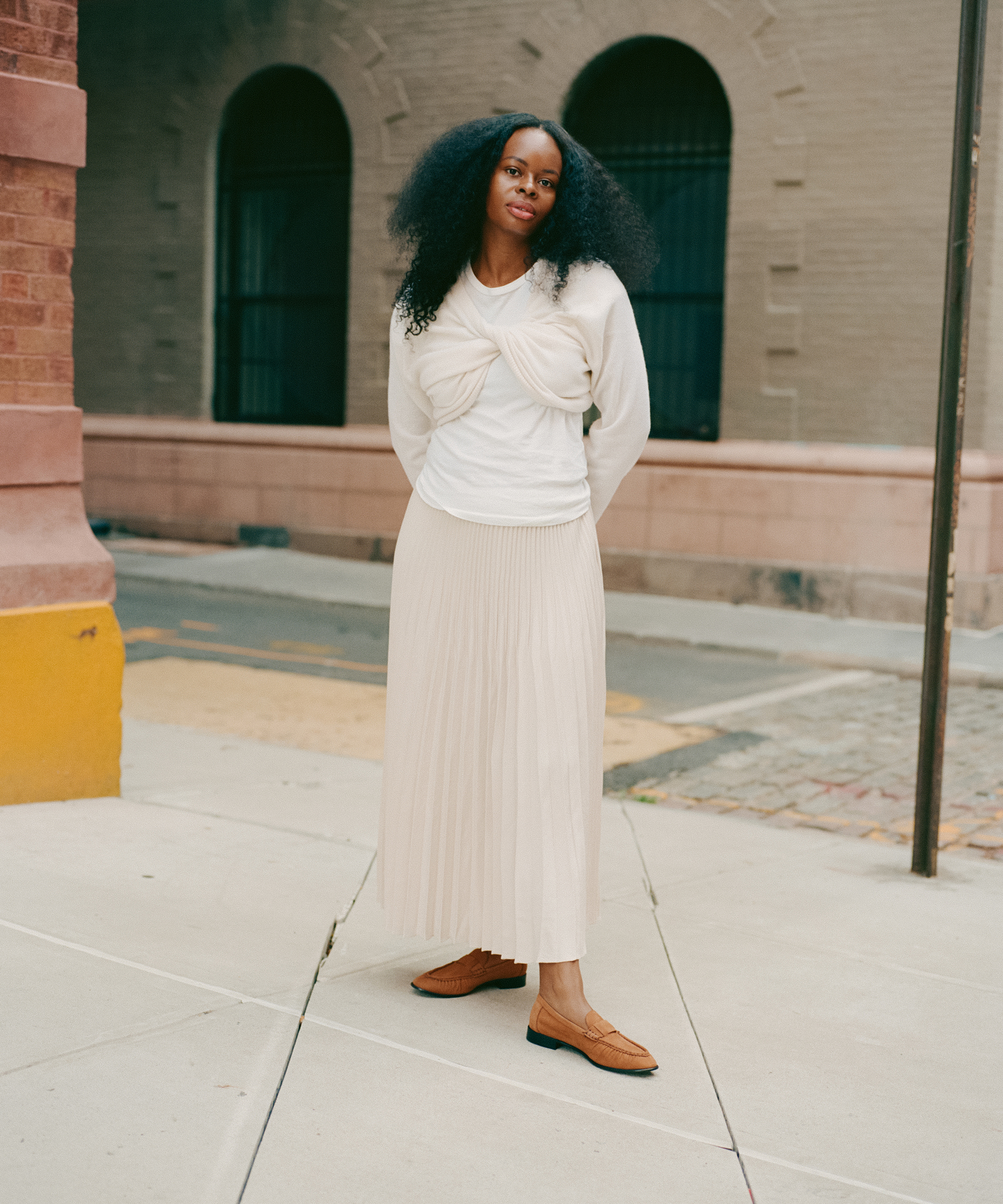 A woman with long, curly hair stands on a city sidewalk wearing a cream long-sleeved top with a twist detail, a matching pleated skirt, and brown shoes, with her hands behind her back. A brick and stone building is in the background.
