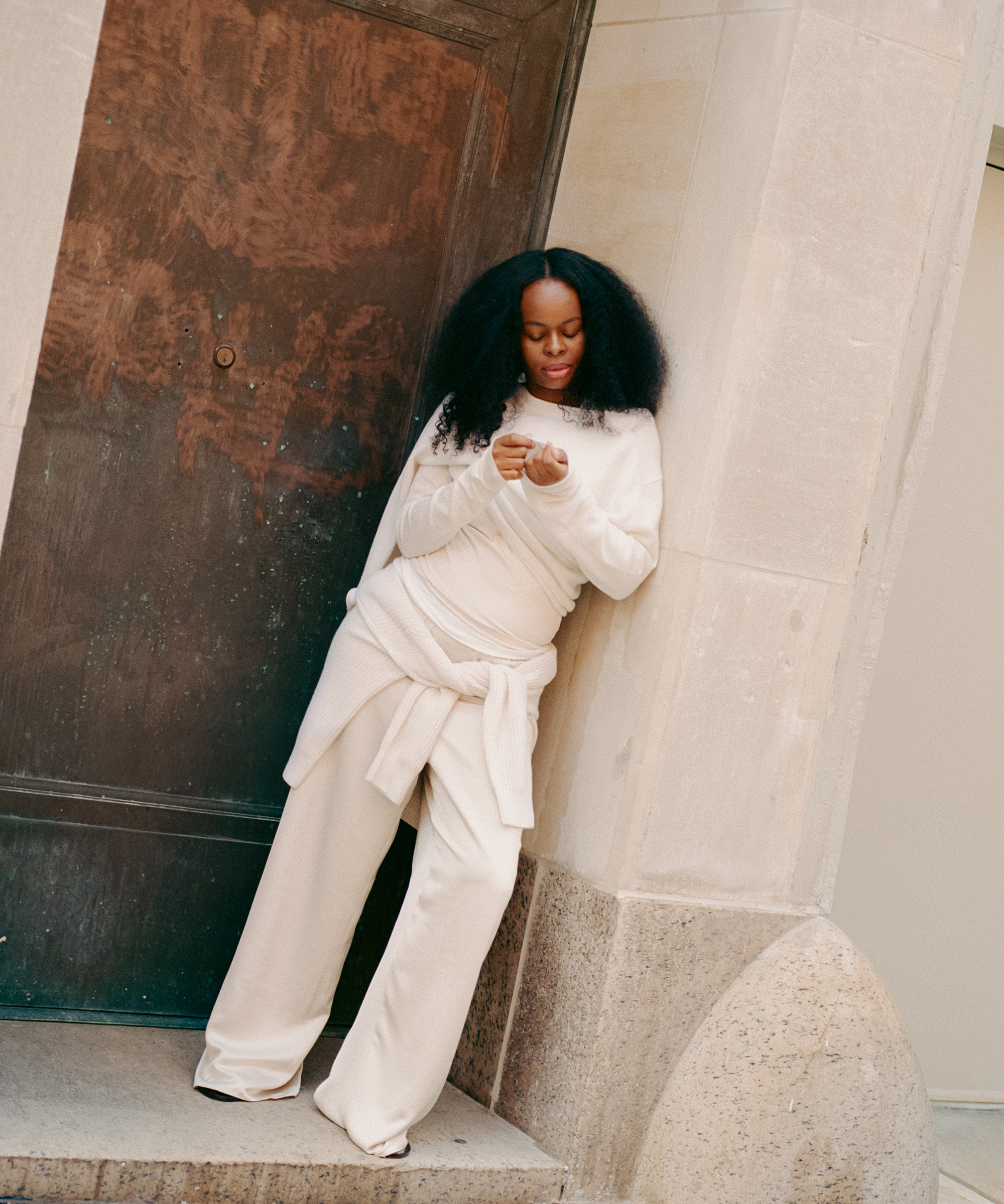 A woman with voluminous curly hair, dressed in a cream-colored outfit with a sweater tied around her waist, stands leaning against a stone doorway, looking down at her hands.