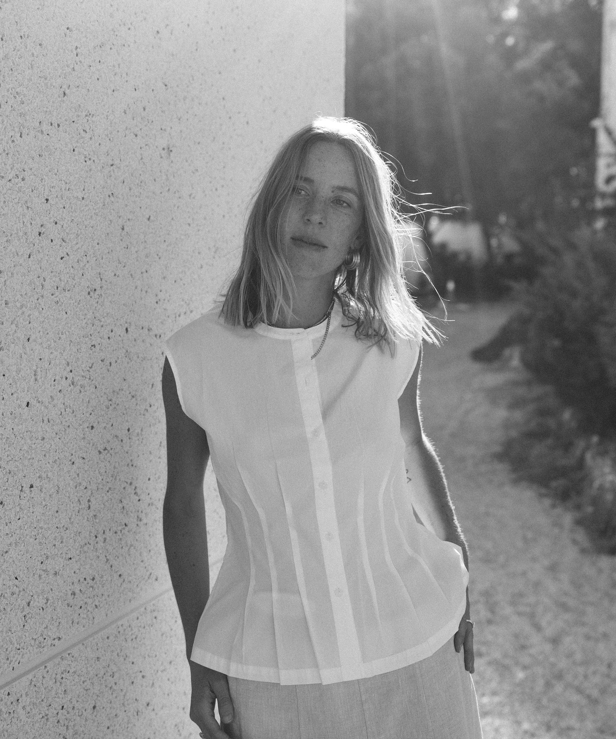 Black-and-white photo of Nuria Val, a woman with light hair in a sleeveless, button-up white top, standing outdoors by a textured wall as sunlight shines from behind her.