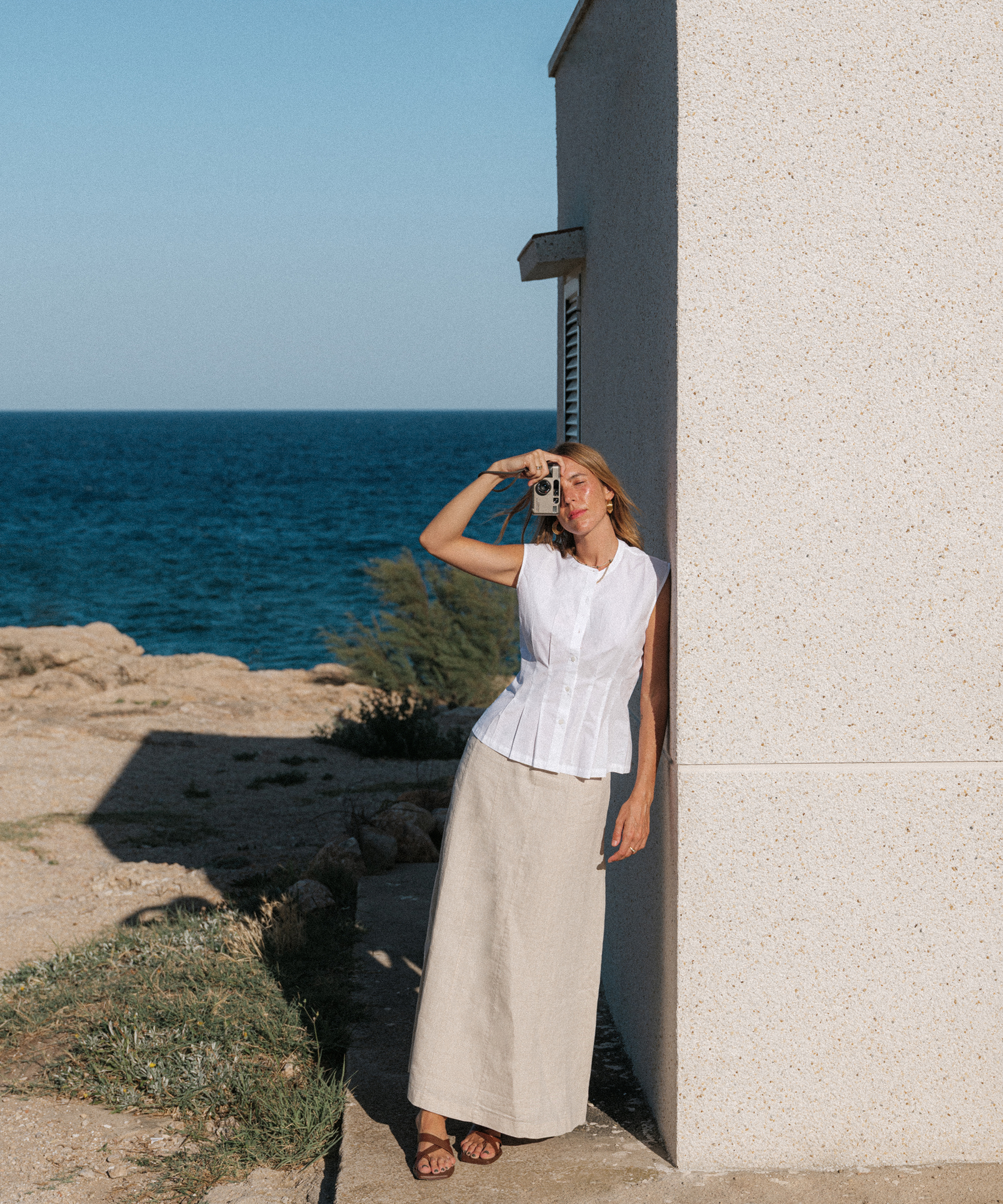 A woman in a white sleeveless top and long beige skirt stands against a white wall by the sea, holding a camera to her eye as she faces the ocean under clear blue skies—capturing the moment in true Nuria Val style.