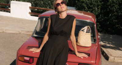 Nuria Val, in a sleeveless black dress and sunglasses, leans back on the hood of a red vintage car. A woven straw bag sits beside her. The sunny outdoor scene is framed by lush greenery in the background.