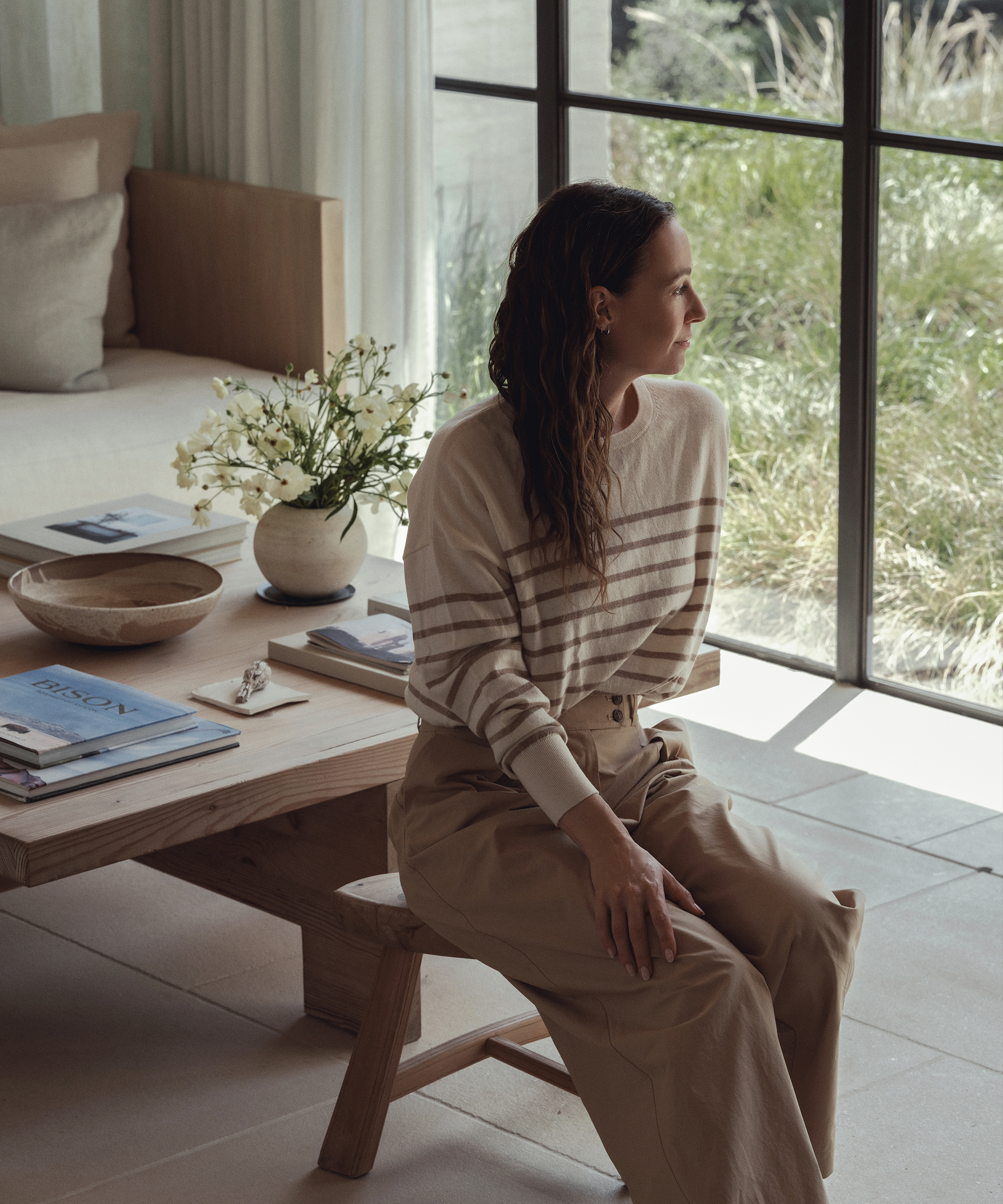 A woman with long brown hair, wearing a striped jenni kayne sweater and beige pants, sits on a wooden bench in a bright, modern living room, gazing out a large window with greenery. A wooden table with books and a vase sits nearby.