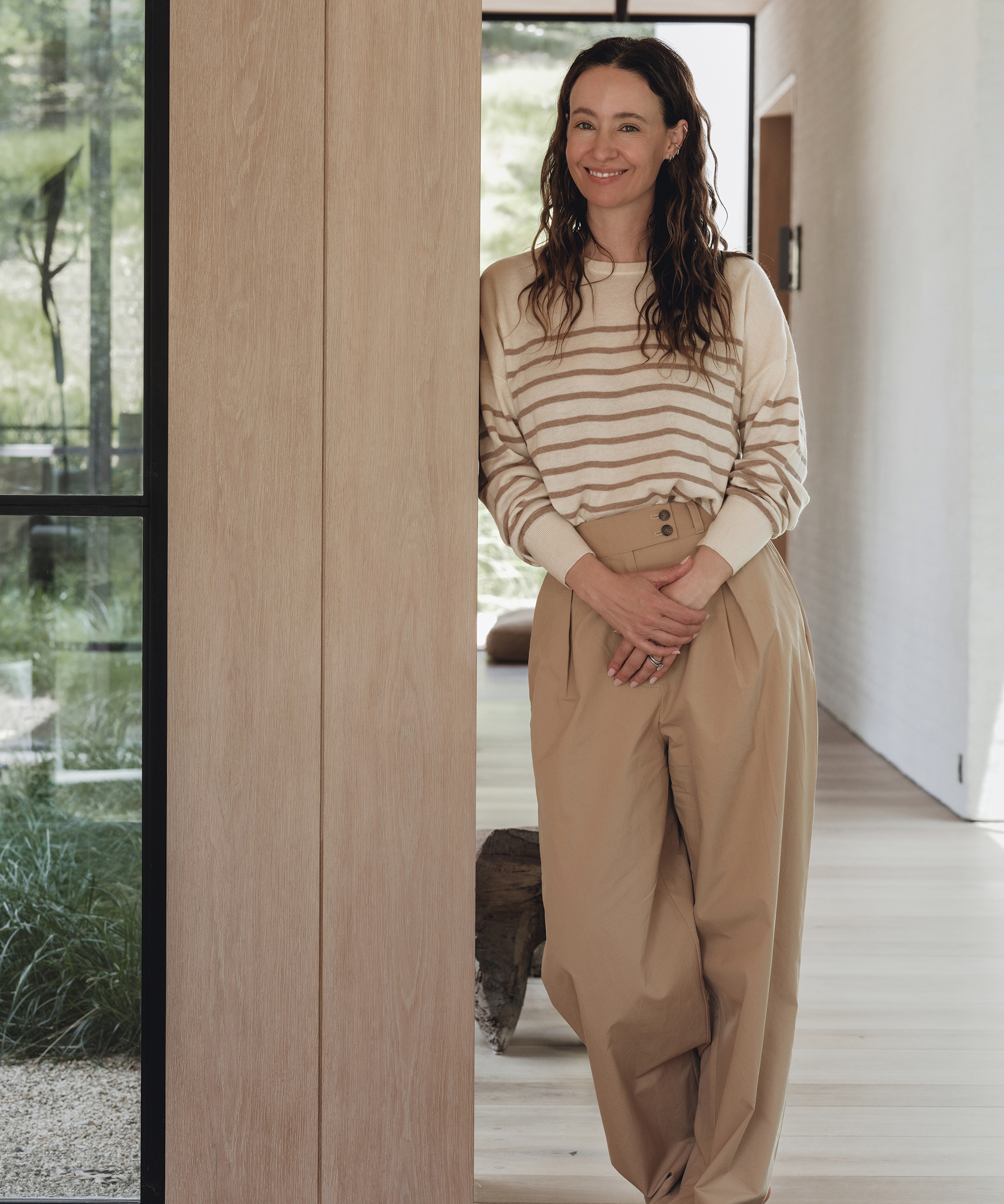 A woman with long wavy hair, wearing a Jenni Kayne cream and beige striped sweater and high-waisted beige trousers, stands indoors by a wooden wall, smiling with her hands clasped as light streams in through large windows.