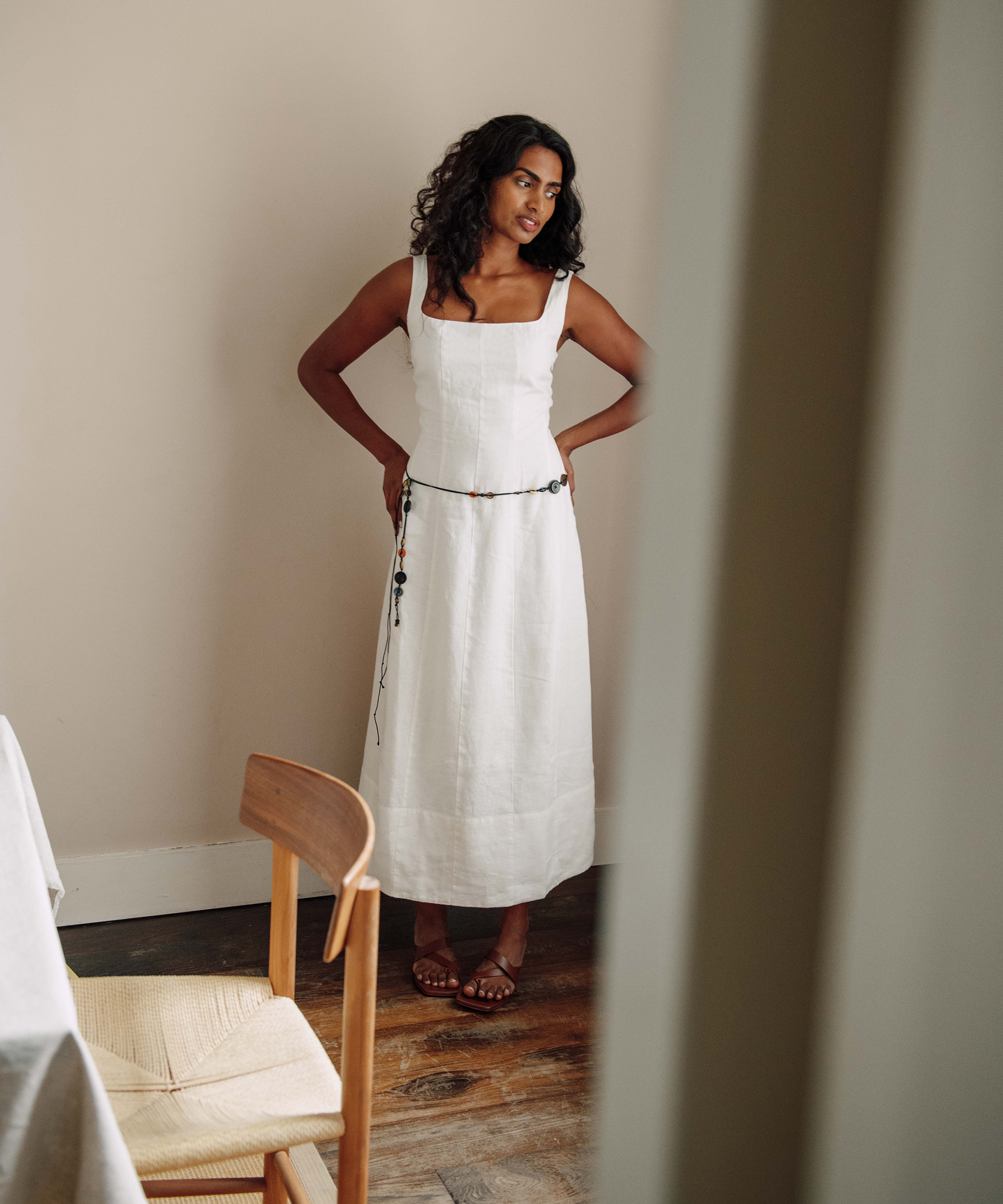 Kirthanaa Naidu, with long curly hair, stands indoors wearing a white sleeveless dress and sandals, hands on her hips. A wooden chair and table are partially visible in the foreground.