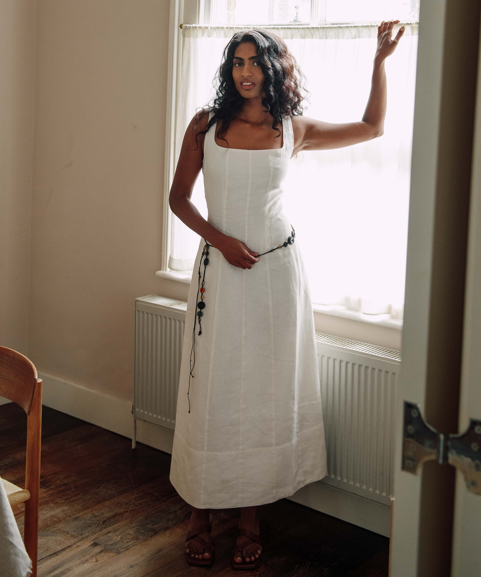 A woman in a long white dress stands by a window, holding the curtain open. She has wavy dark hair, wears sandals, and stands on a wooden floor in a softly lit room with cream-colored walls, reminiscent of Kirthanaa Naidu’s serene style.