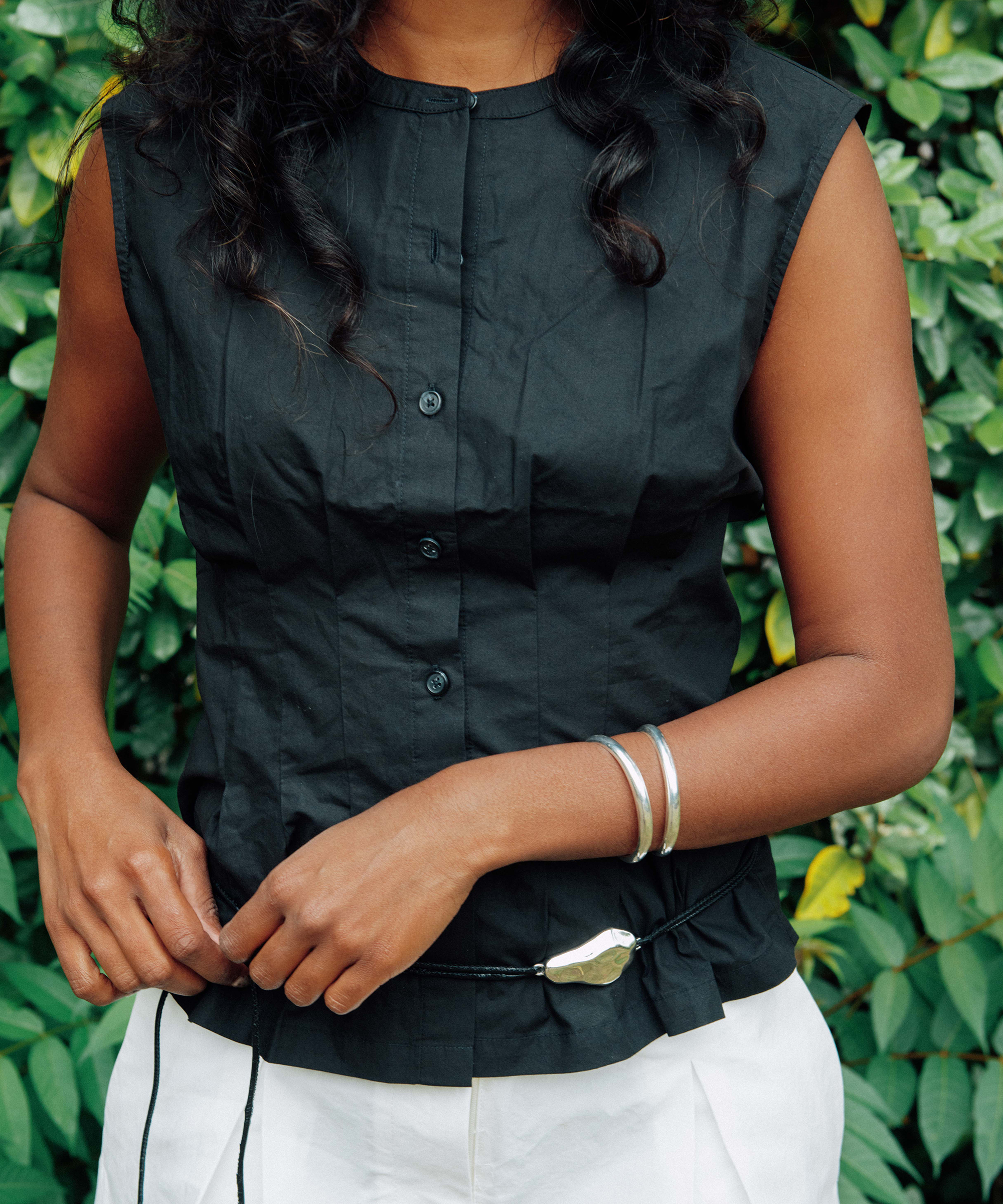 A person with wavy hair, possibly Kirthanaa Naidu, wears a sleeveless black button-up shirt, white pants, and silver bracelets, standing in front of green leafy plants. Only their torso and hands are visible.