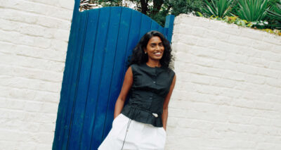 A woman with long curly hair, wearing a black sleeveless top and white skirt, stands smiling in front of a bright blue wooden gate and a white brick wall. Green plants are visible above the wall—Kirthanaa Naidu radiates joy in this vibrant setting.
