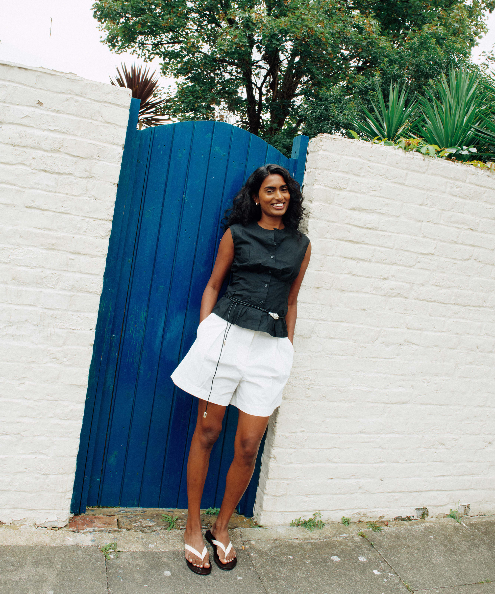 Kirthanaa Naidu stands smiling in front of a white brick wall and blue wooden gate, wearing a black sleeveless top, white shorts, and flip-flops. Green plants and trees are visible behind the wall.