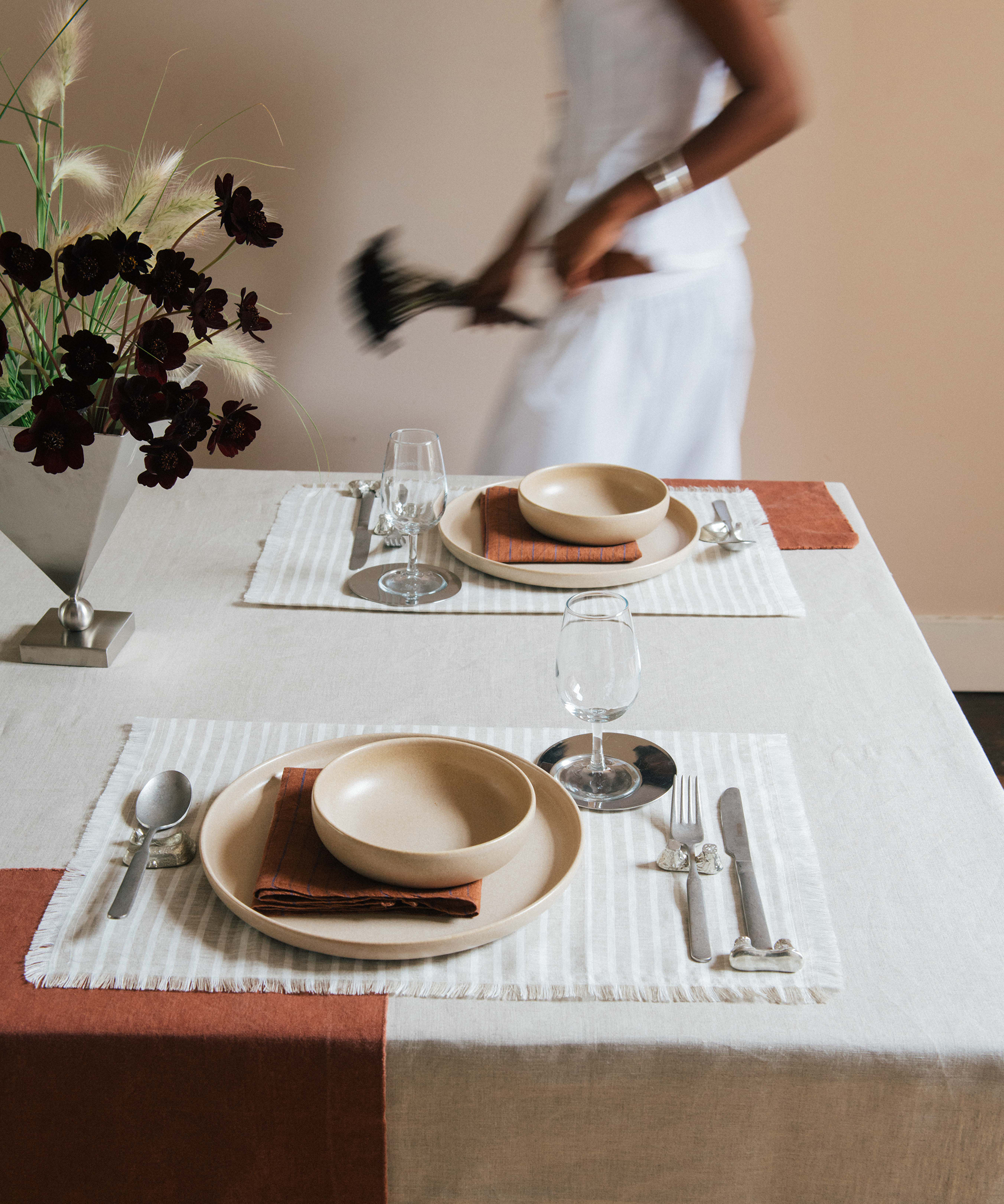 Kirthanna Naidu walking past a table set for two with beige plates, brown napkins, glasses, and silverware on a white and rust-colored tablecloth. A person in white moves behind the table, holding a dark object. A vase with dark flowers is on the table.
