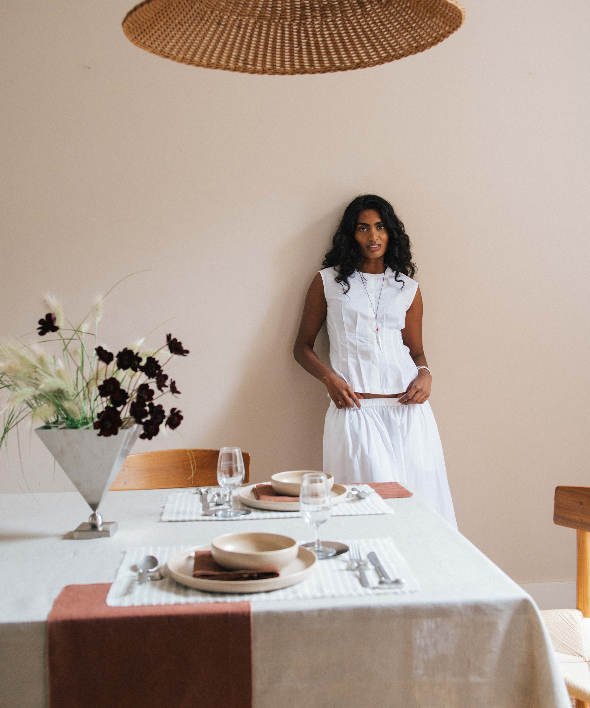 A woman in a sleeveless white dress stands by a beige wall near a dining table set with plates, glasses, and napkins. A woven light fixture hangs above and a vase of flowers decorates the table, styled by Kirthanaa Naidu.