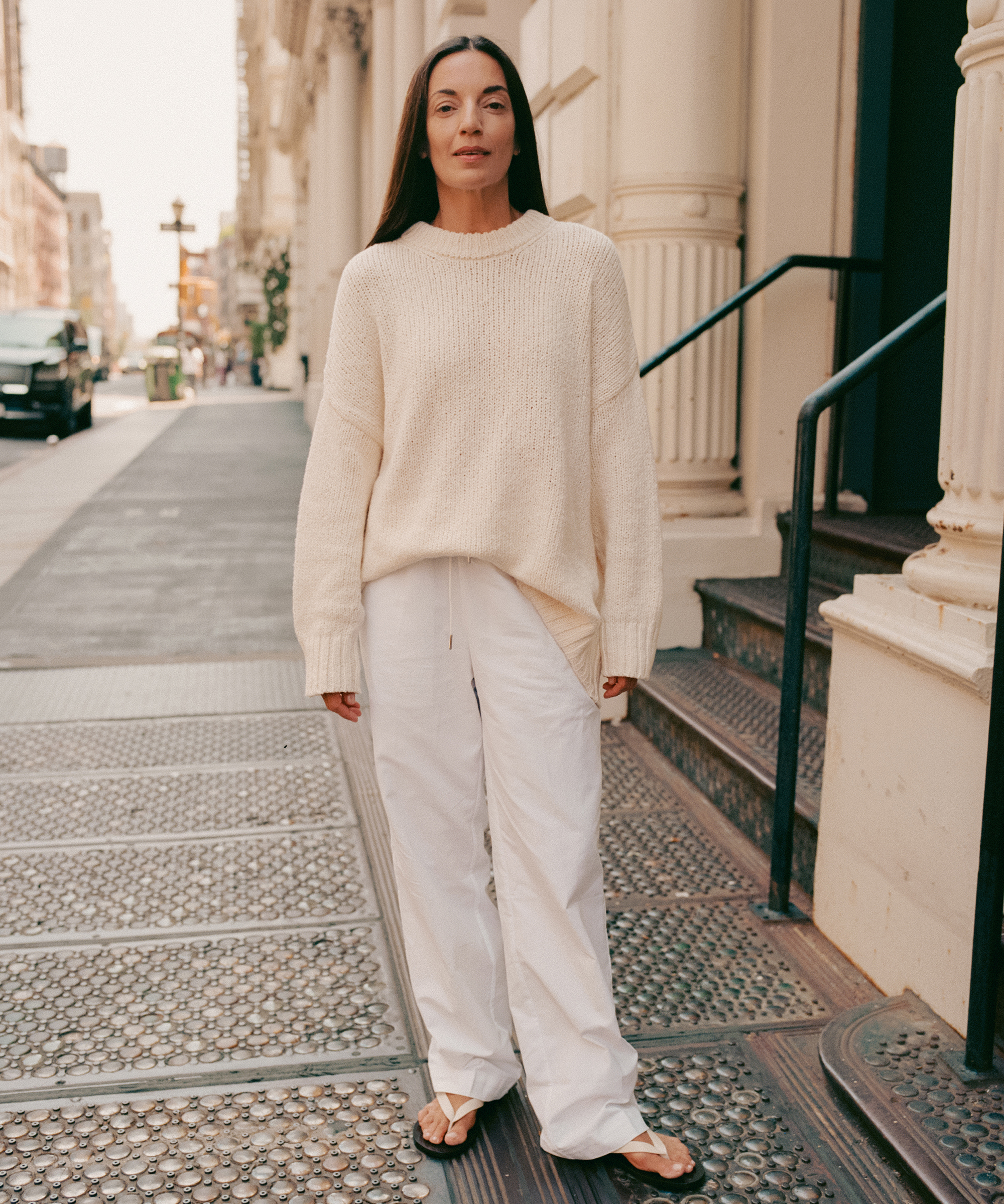 A woman stands on a city sidewalk wearing a cream knit sweater, loose white pants, and sandals. She has long straight hair and a neutral expression, with urban buildings and a touch of metaflora visible in the background.