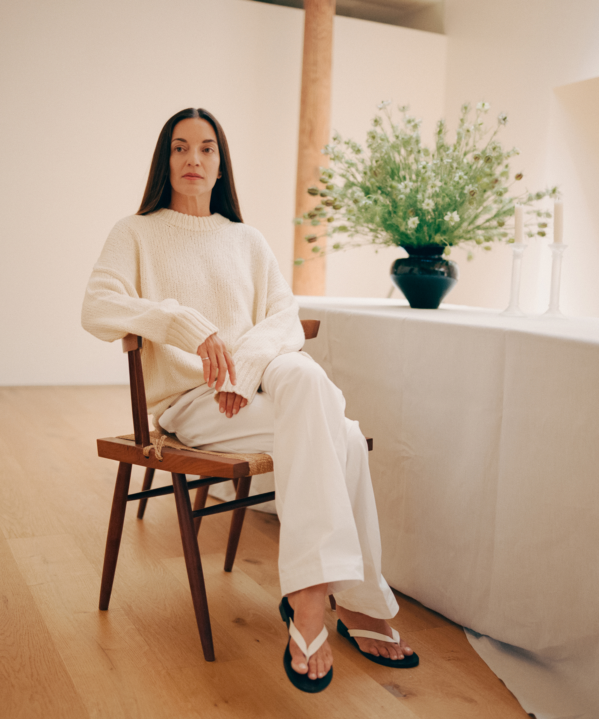 A woman with long dark hair sits on a wooden chair indoors, wearing a cream sweater, white pants, and sandals. She is near a table with a white tablecloth, two white candlesticks, and a metaflora-inspired black vase of flowers.