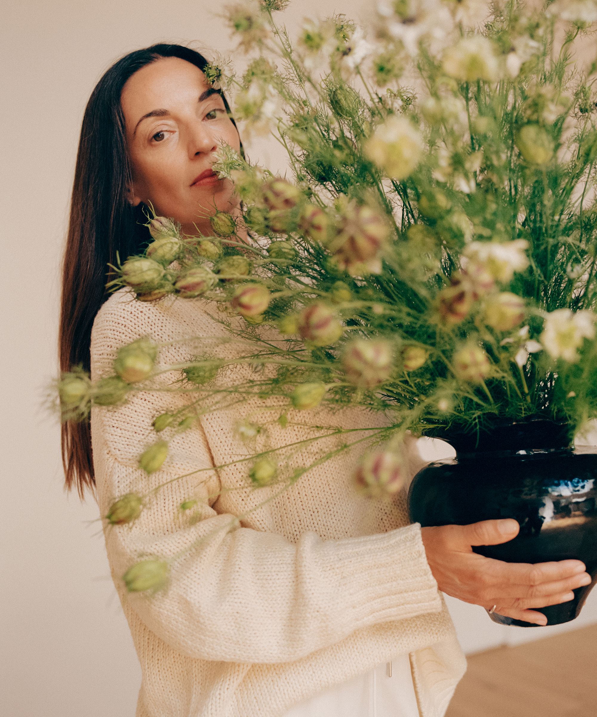 A woman in a cream sweater holds a large black metaflora vase filled with green, leafy flowers, partially obscuring her face. She stands indoors against a neutral background.