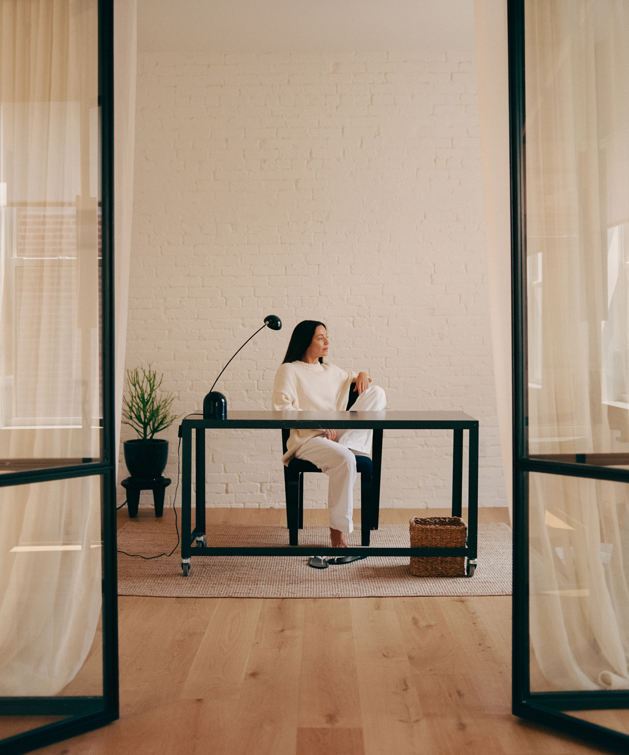 A woman dressed in white sits at a modern black desk in a minimalist room with white brick walls, a metaflora plant, a desk lamp, and a woven basket on the floor. Natural light fills the space, creating a calm atmosphere.