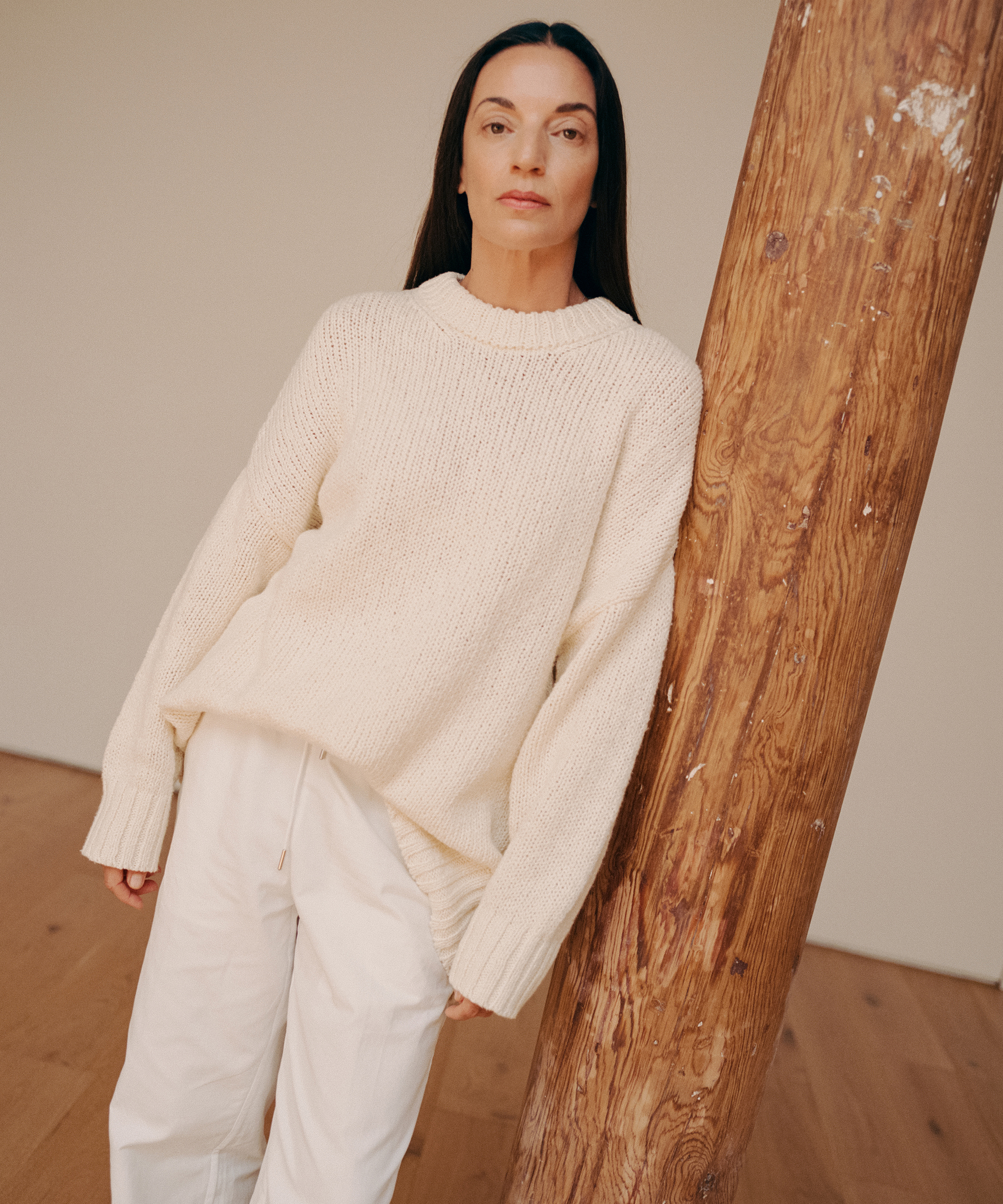 A woman with long dark hair, wearing a cream-colored sweater and white pants, stands indoors leaning against a wooden pillar on a light wood floor, looking directly at the camera in a serene, metaflora-inspired setting.